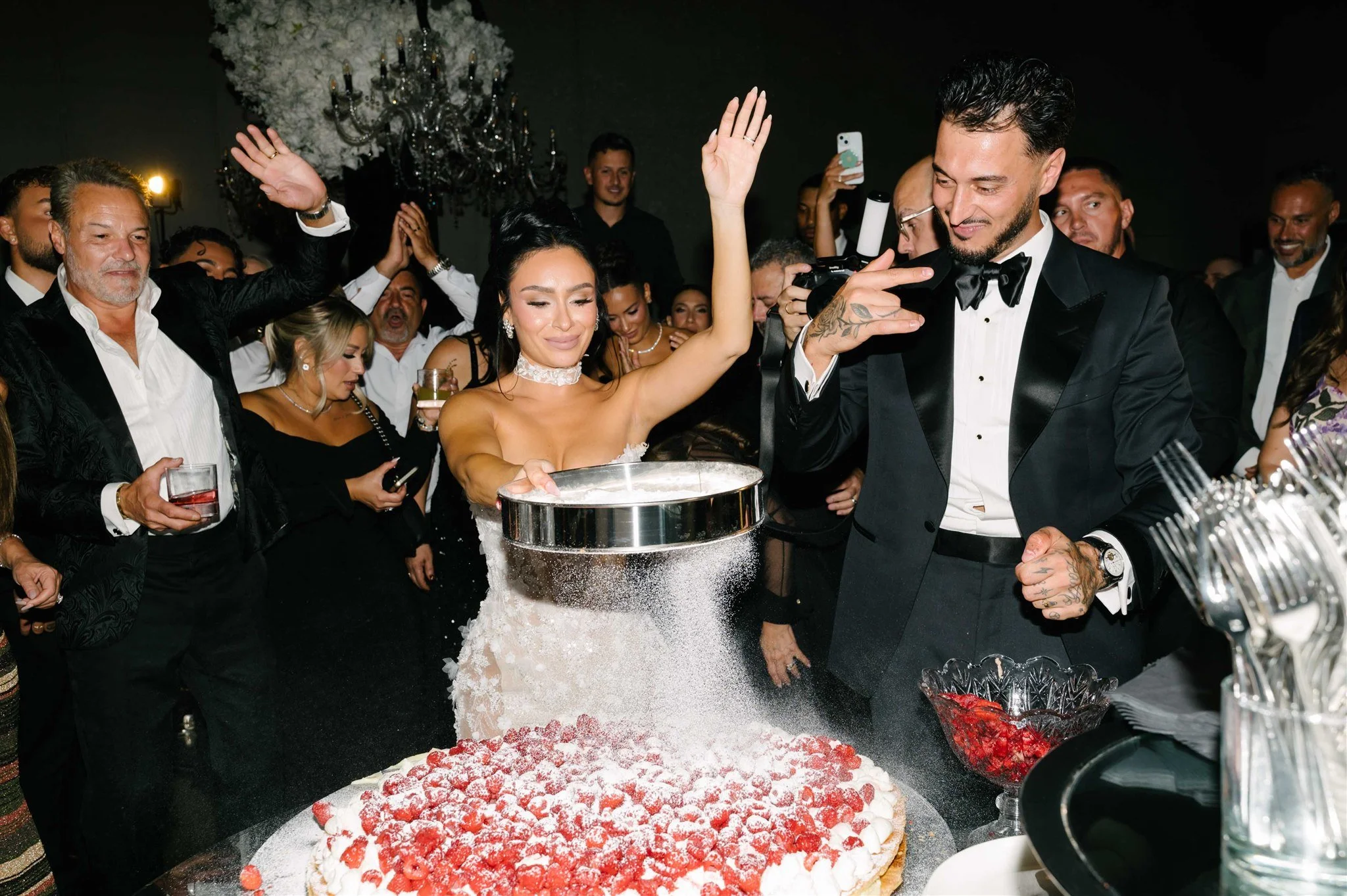 A wedding celebration where the bride and groom are participating in a cake slicing tradition. The bride is smiling and holding a cake knife, while the groom is pointing and looking at her. They are surrounded by guests in formal attire, with some ca