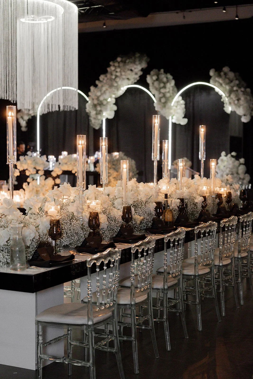 Elegant event table with tall candles, white floral arrangements, and clear chairs, decorated with arches and floral wall accents.