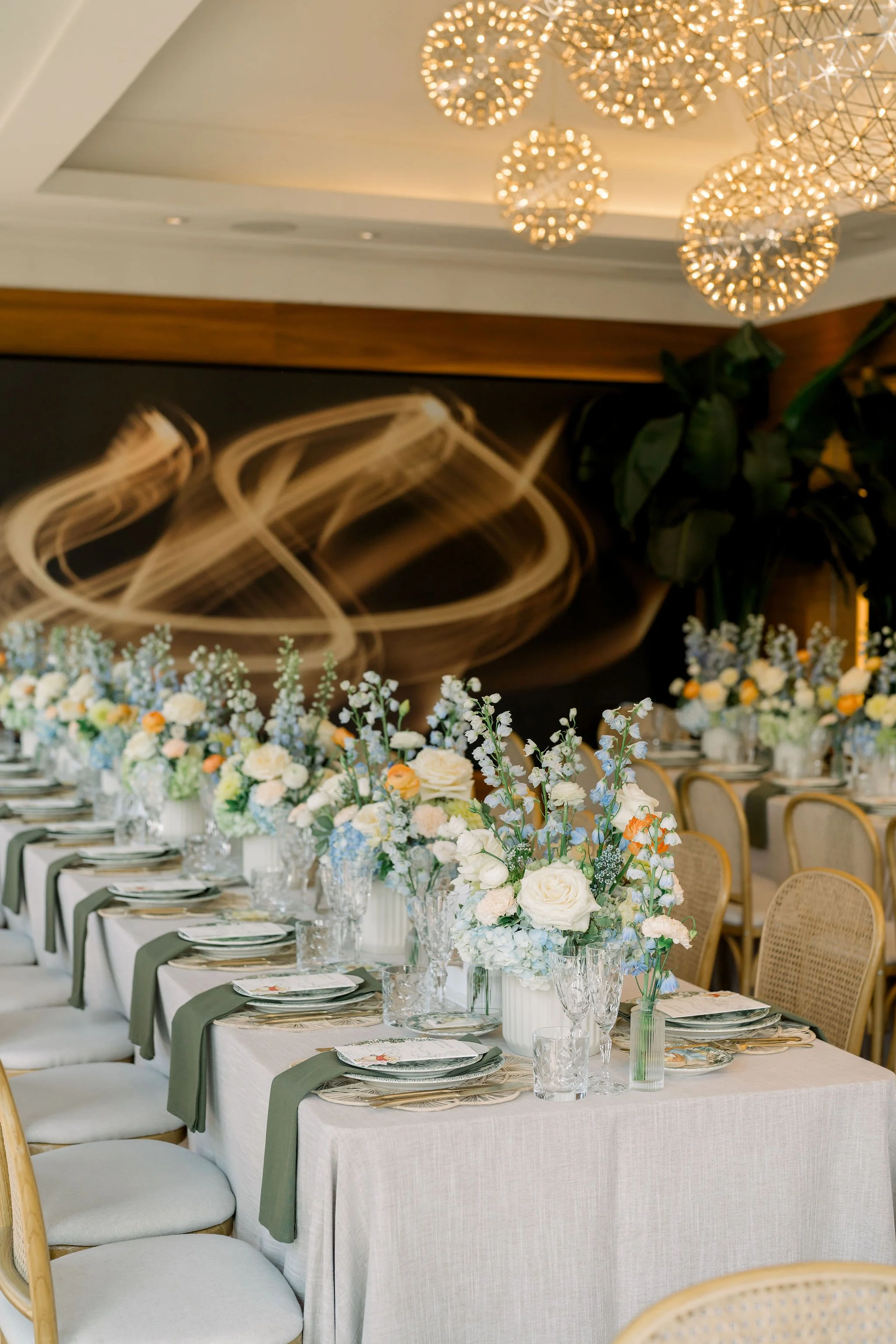 Long banquet table decorated with white tablecloths, floral centerpieces, and place settings, set in a luxurious indoor venue with modern chandeliers and greenery.