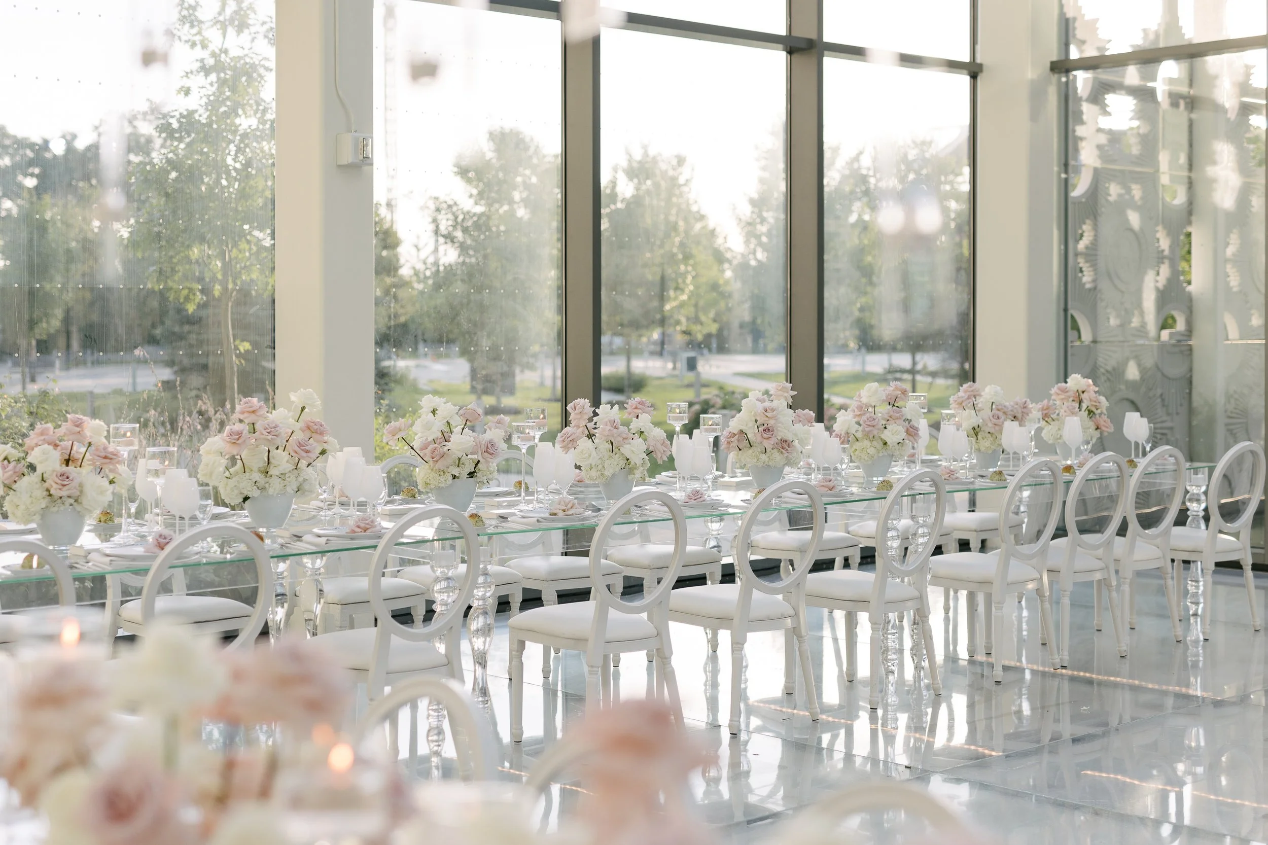 Elegant banquet table setting with white chairs, floral centerpieces, and glassware inside a modern venue with floor-to-ceiling windows overlooking outdoor greenery.