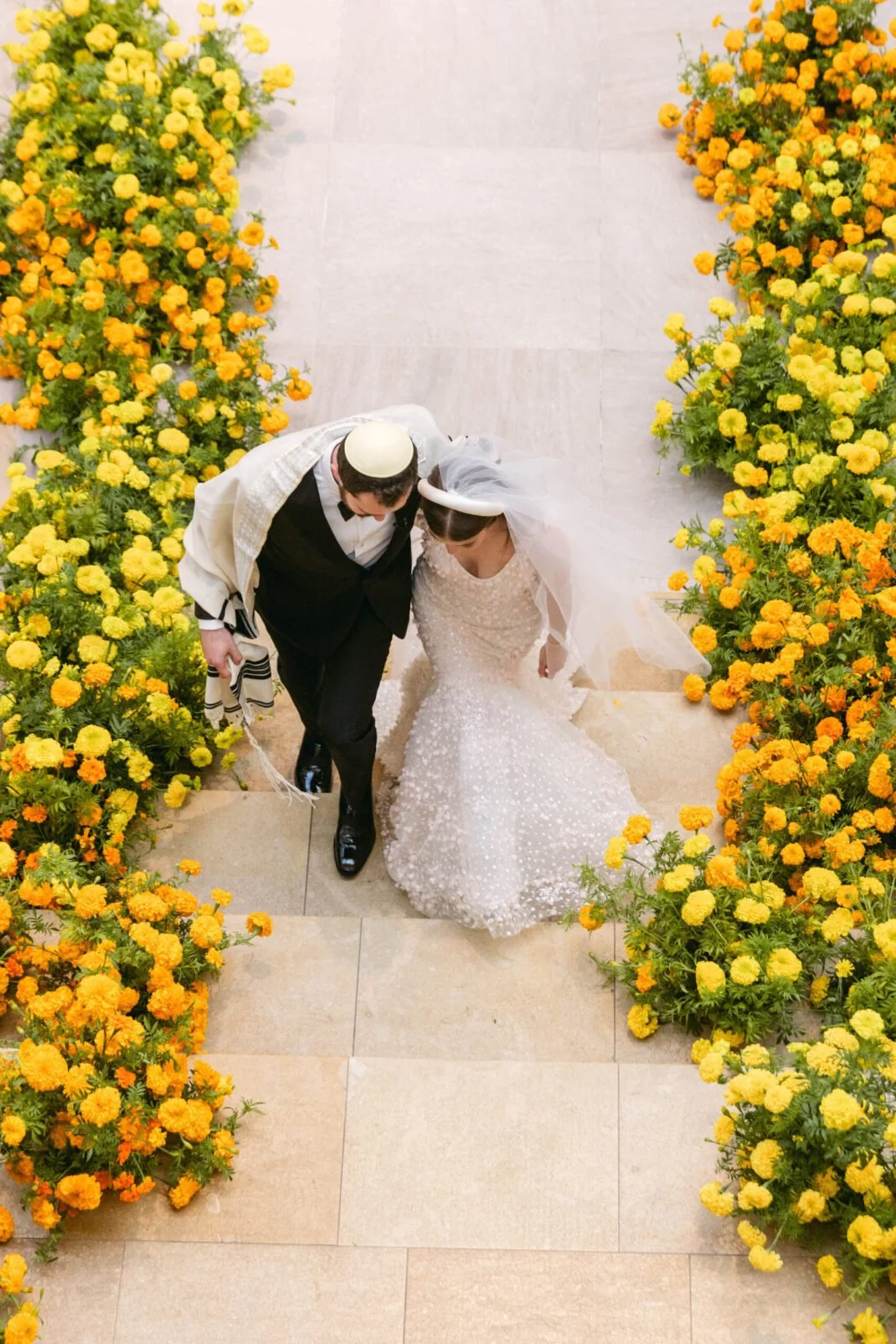 A bride and groom walking up stairs decorated with yellow and orange flowers at a wedding ceremony.