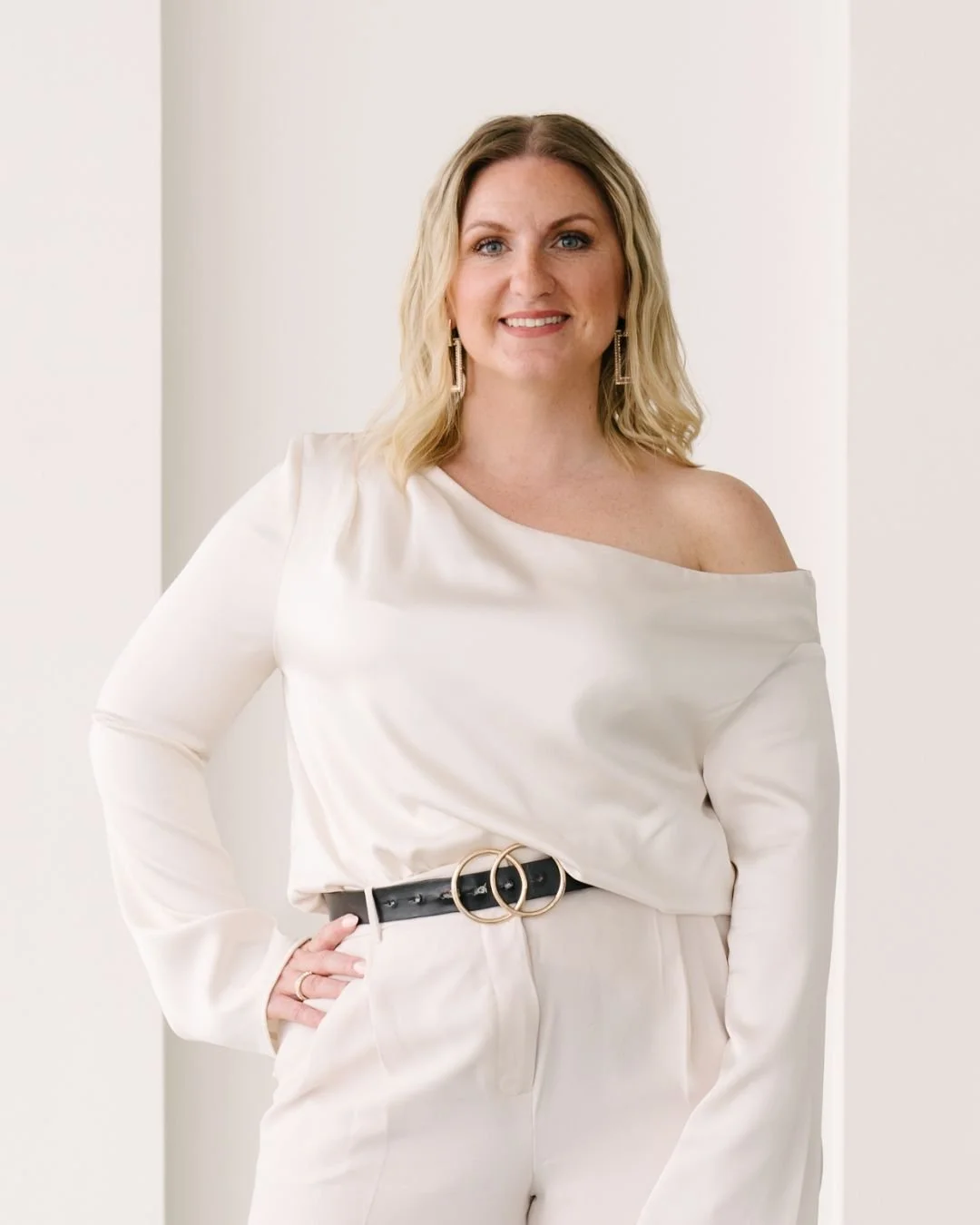 A woman with blonde hair and earrings is smiling, wearing an off-shoulder white top and white pants with a black belt, standing indoors against a plain white wall.