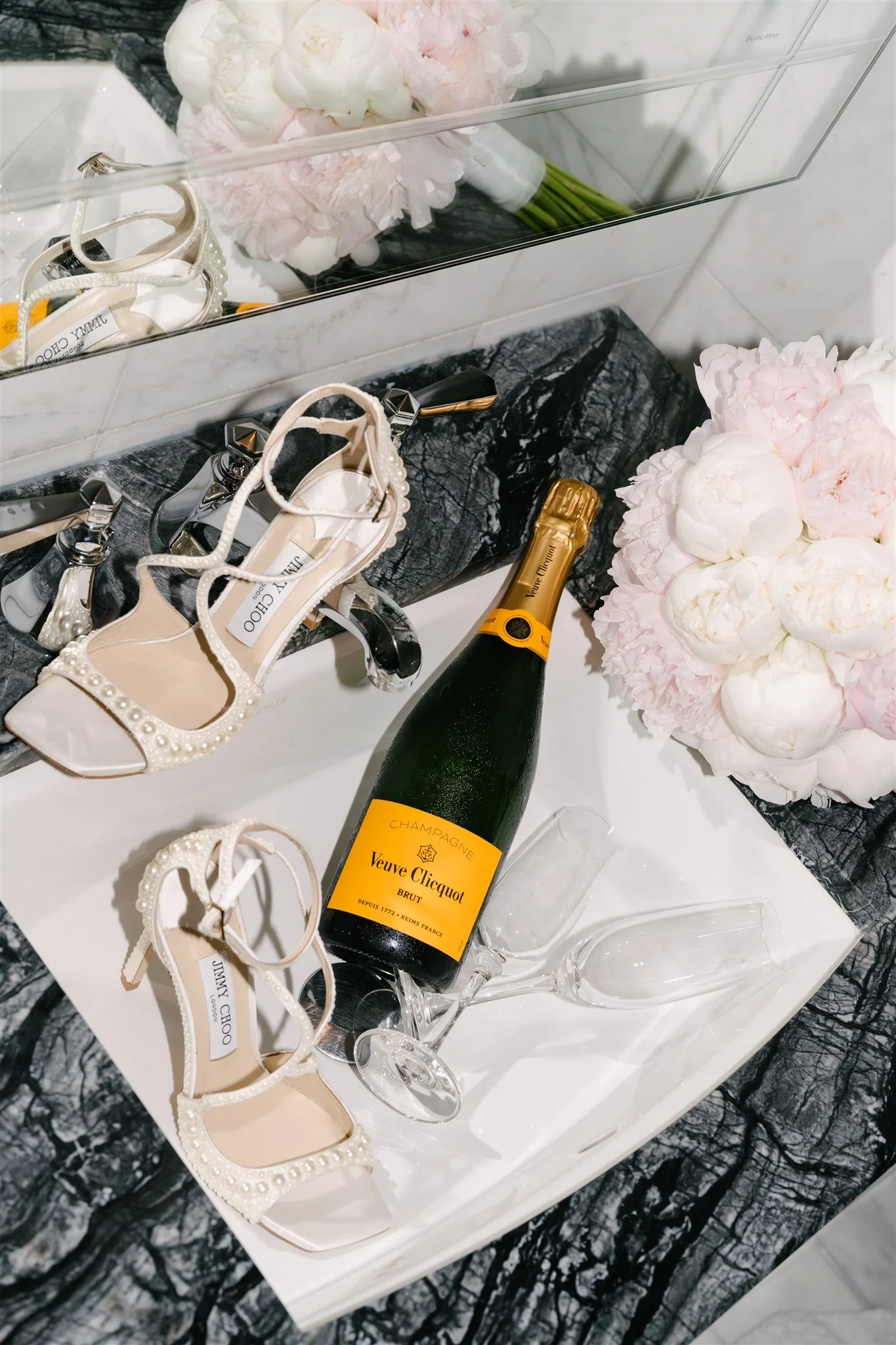 A white rectangular table holding a champagne bottle, empty glasses, a pair of white Jimmy Choo heels decorated with pearls, and pink peonies in the background. The table is on a black and white marble floor with a mirror behind it.