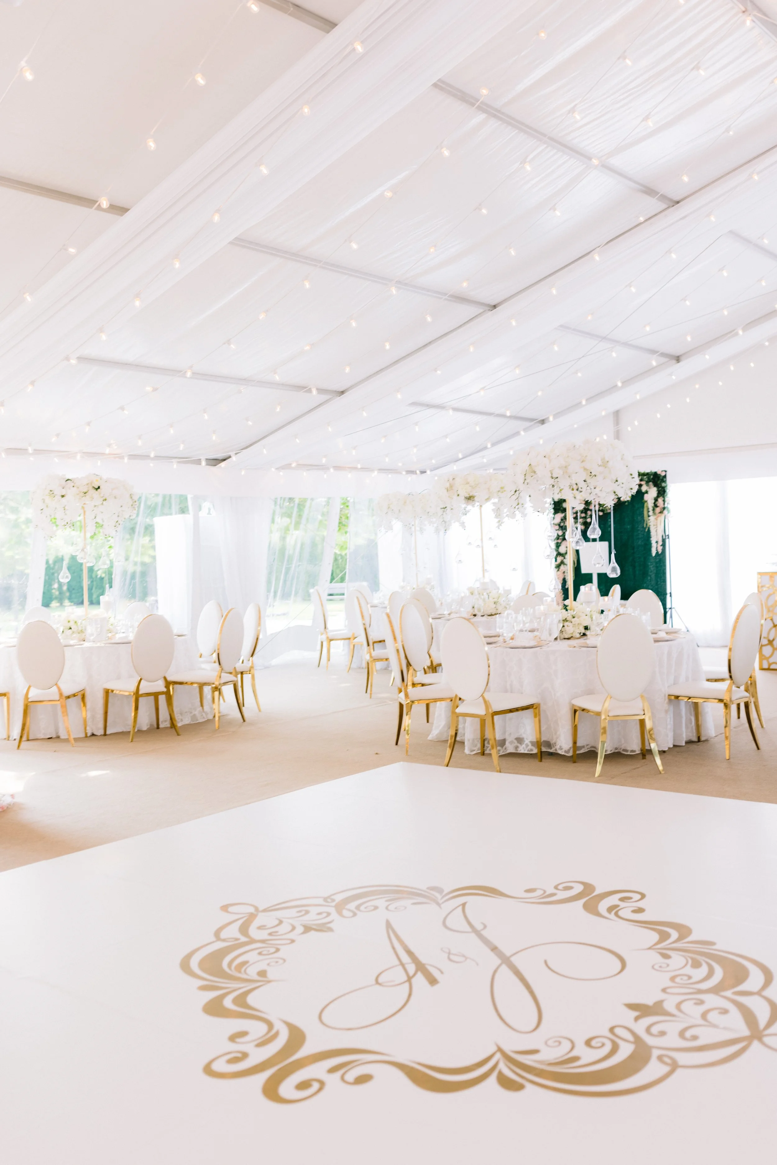 Elegant indoor wedding reception area with round tables covered in white tablecloths, white chairs with gold accents, tall floral centerpieces, and a decorated wedding monogram on the floor.