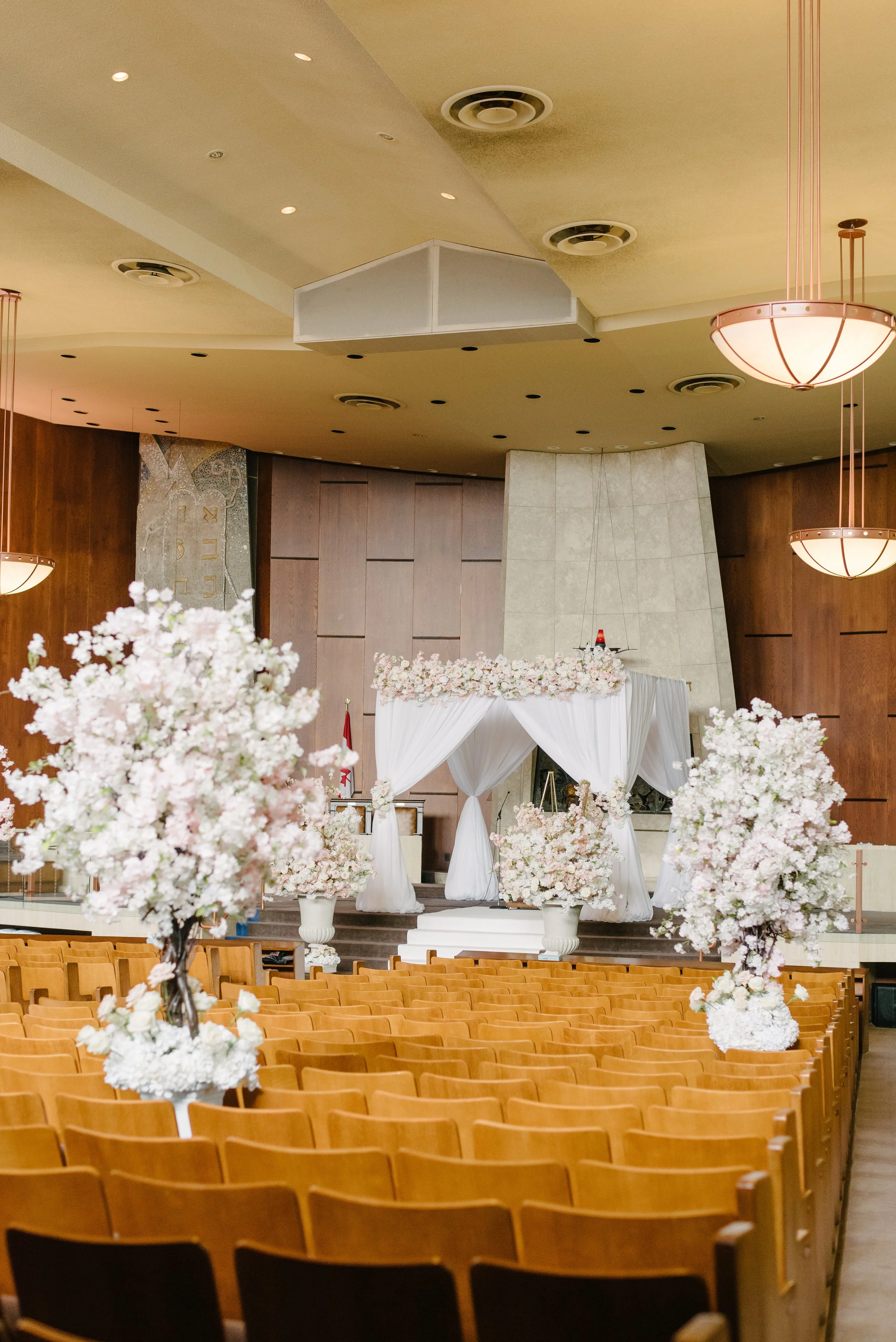 Wedding ceremony setup with an altar adorned with white drapes and pink floral arrangements, surrounded by empty wooden chairs, in a spacious hall with ceiling lights and chandeliers.