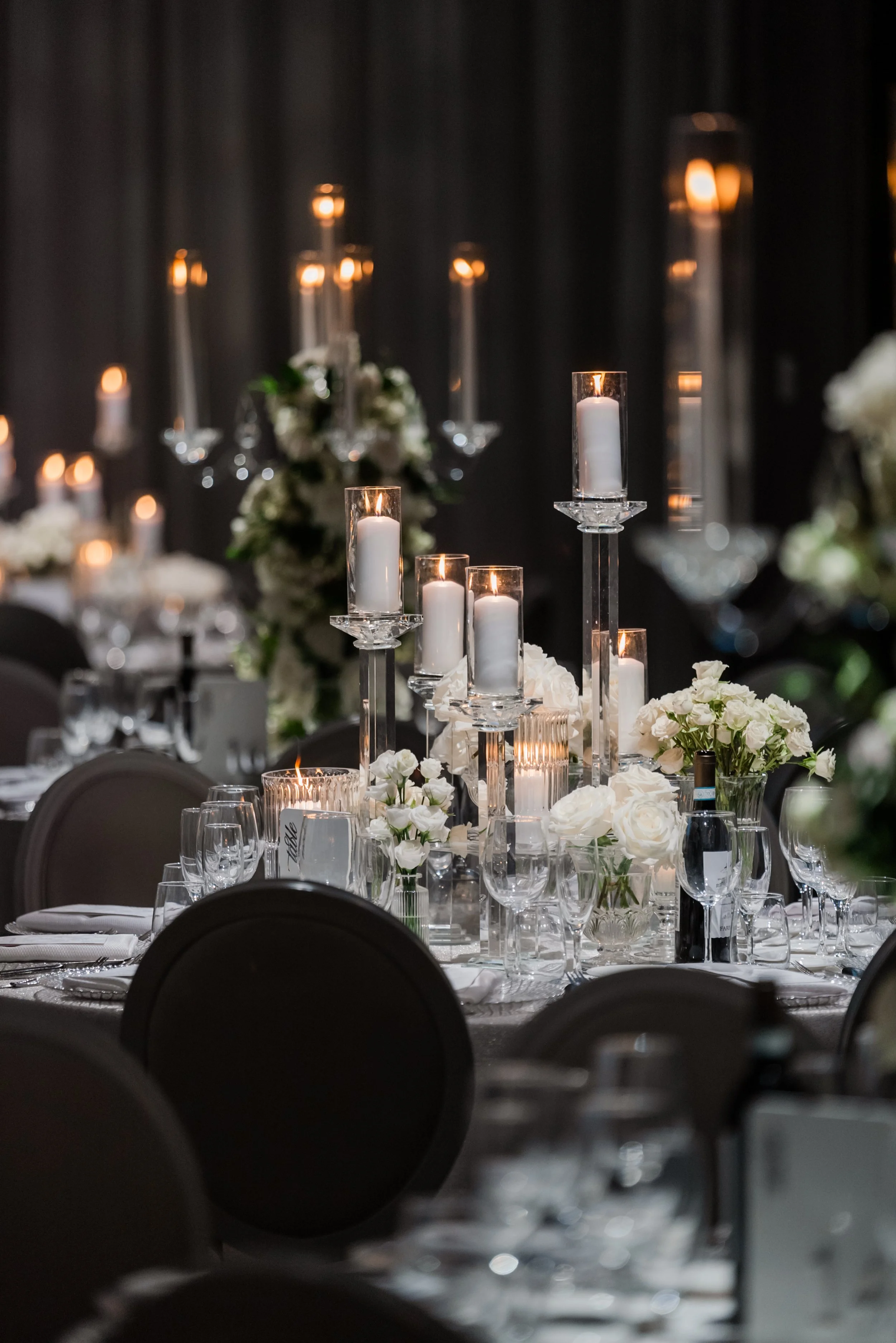 Elegant banquet table with white floral centerpieces and tall glass candle holders with white candles, dimly lit for a formal event, with a dark curtain backdrop.