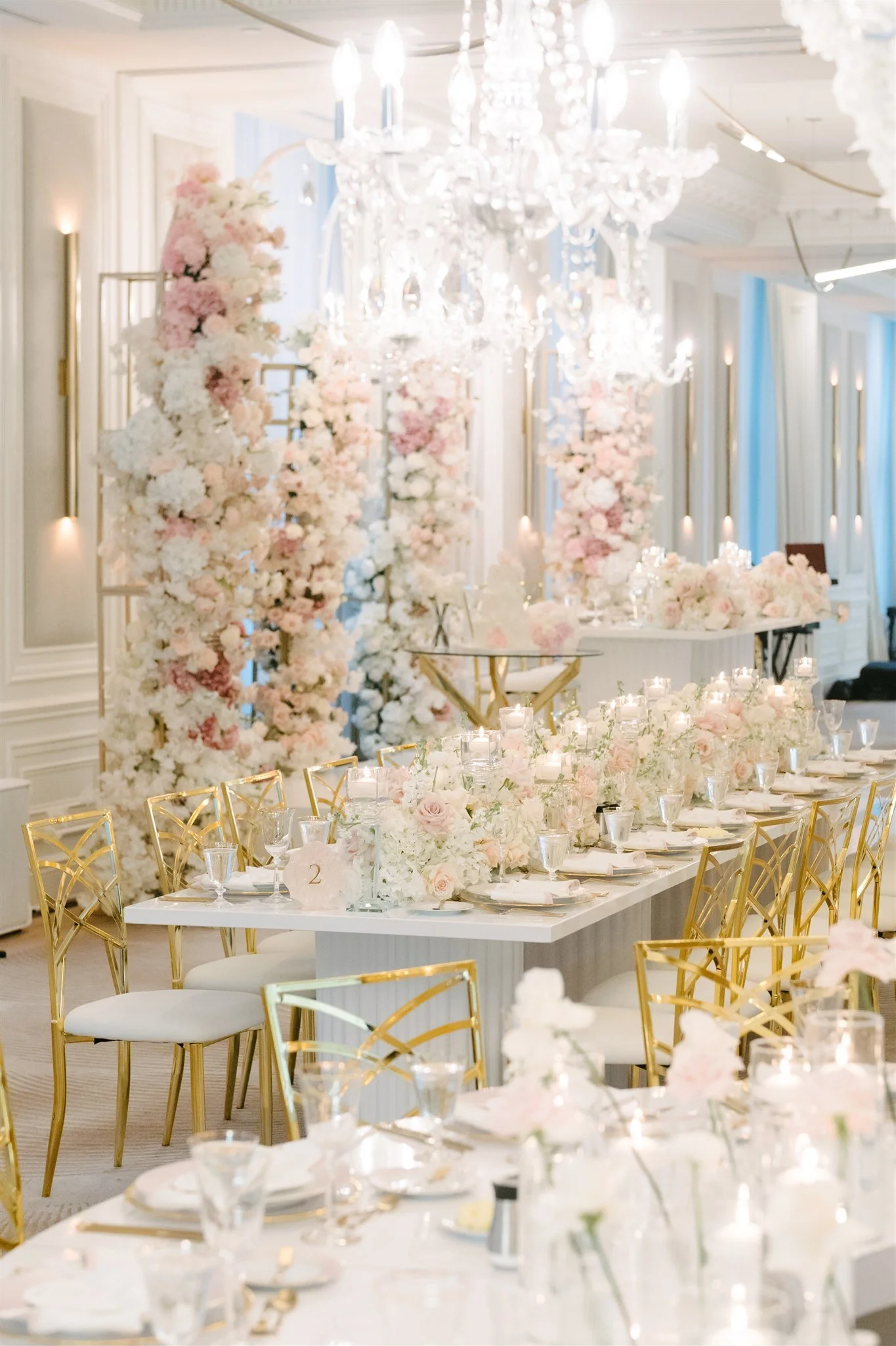Elegant event or wedding reception decorated with floral arrangements, a large chandelier, and gold accents on chairs and tableware.