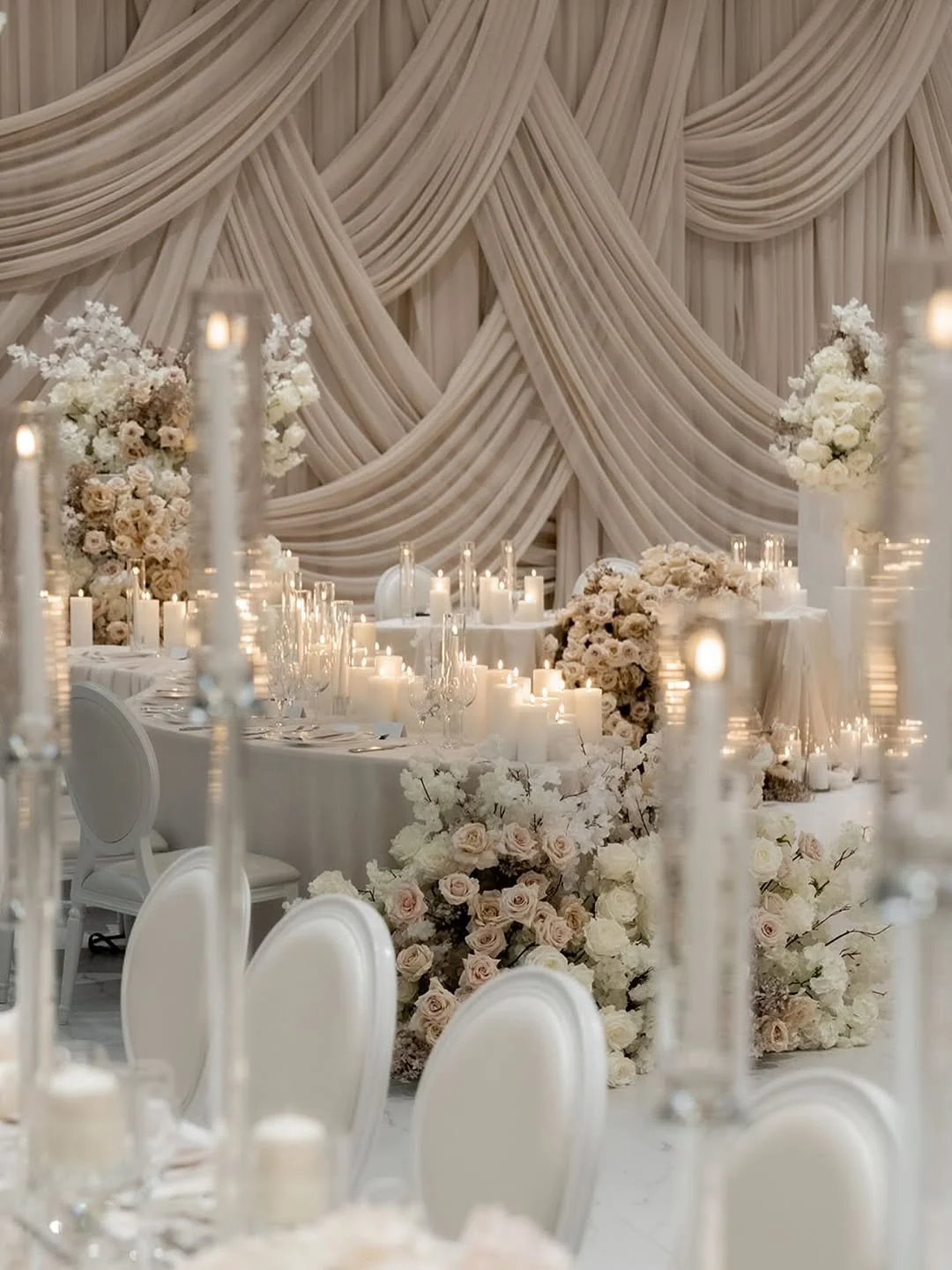 Elegant wedding reception table decorated with white flowers, candles, and glassware, with draped fabric backdrop.