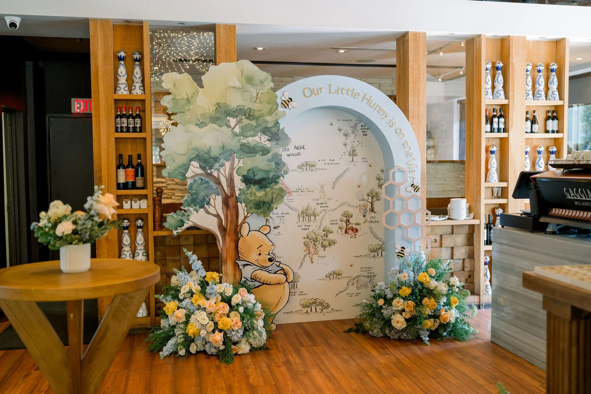 Decorative display featuring a large illustrated tree and Winnie the Pooh character, surrounded by floral arrangements and bee decorations, with a sign reading 'Our Little Honey is on the Way'.