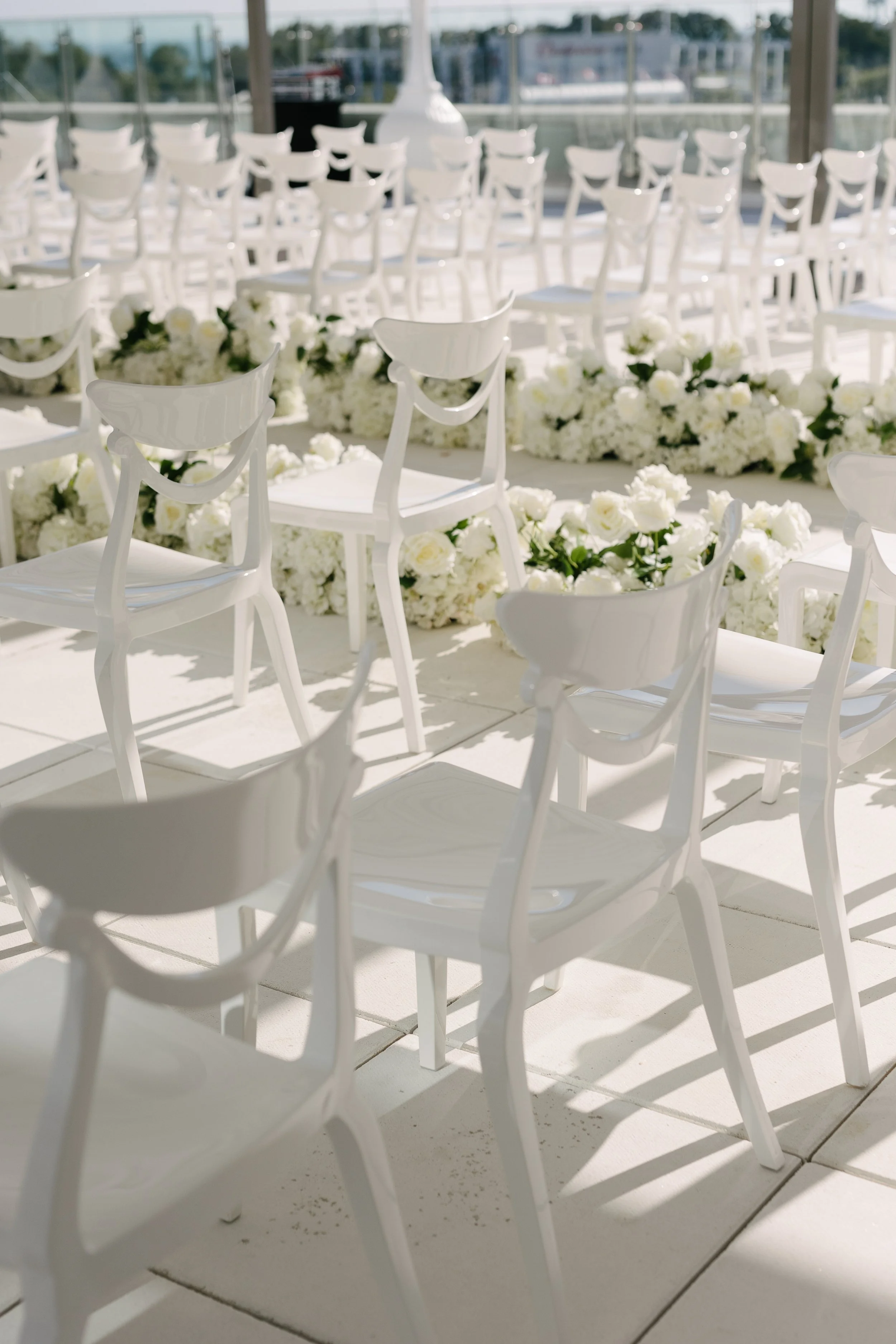 White chairs arranged for a wedding ceremony with white floral decorations on a white floor, set indoors with a glass wall allowing natural light.