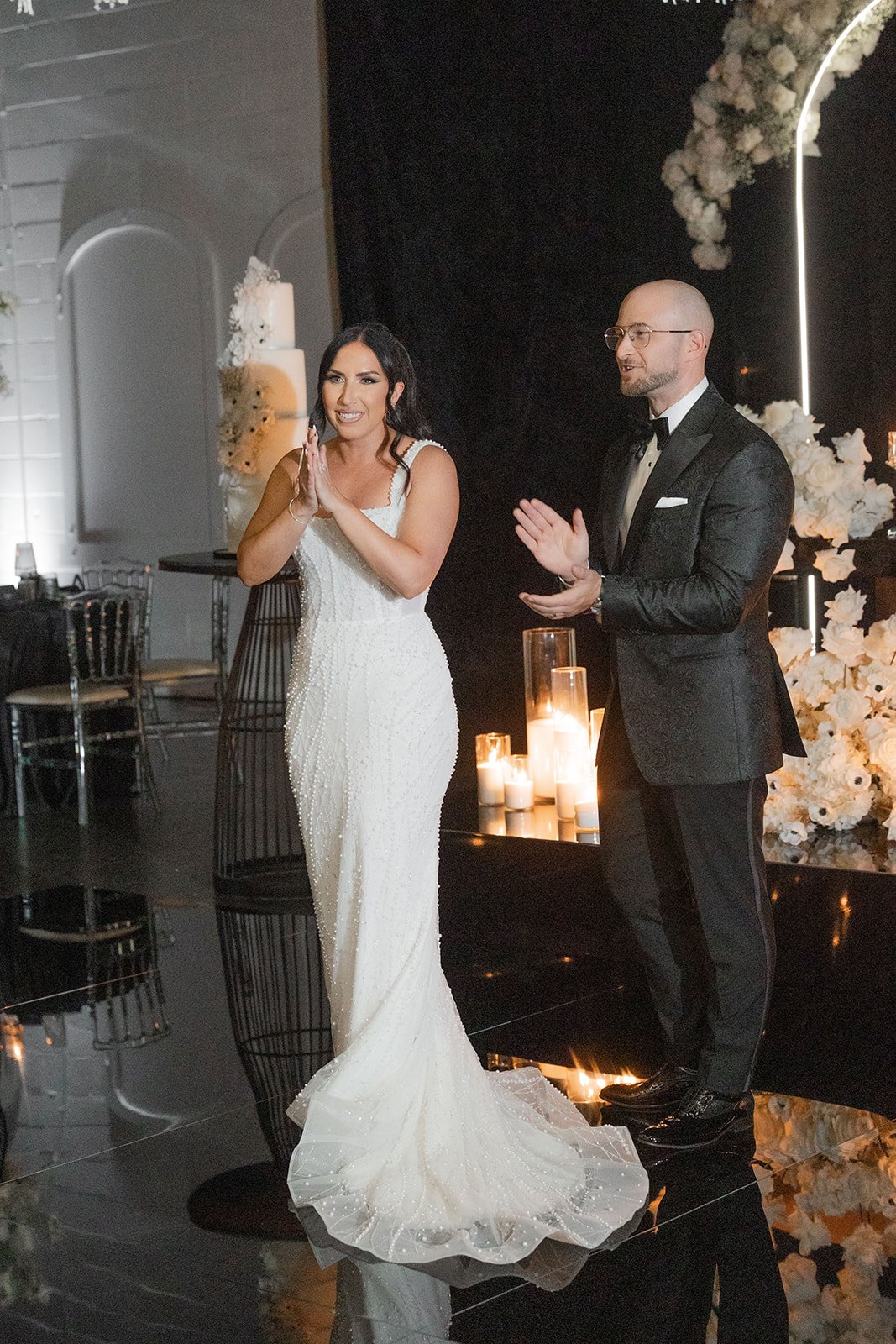 A bride in a white wedding gown standing beside a groom in a black tuxedo, indoors with candles and floral decorations, during a wedding celebration.