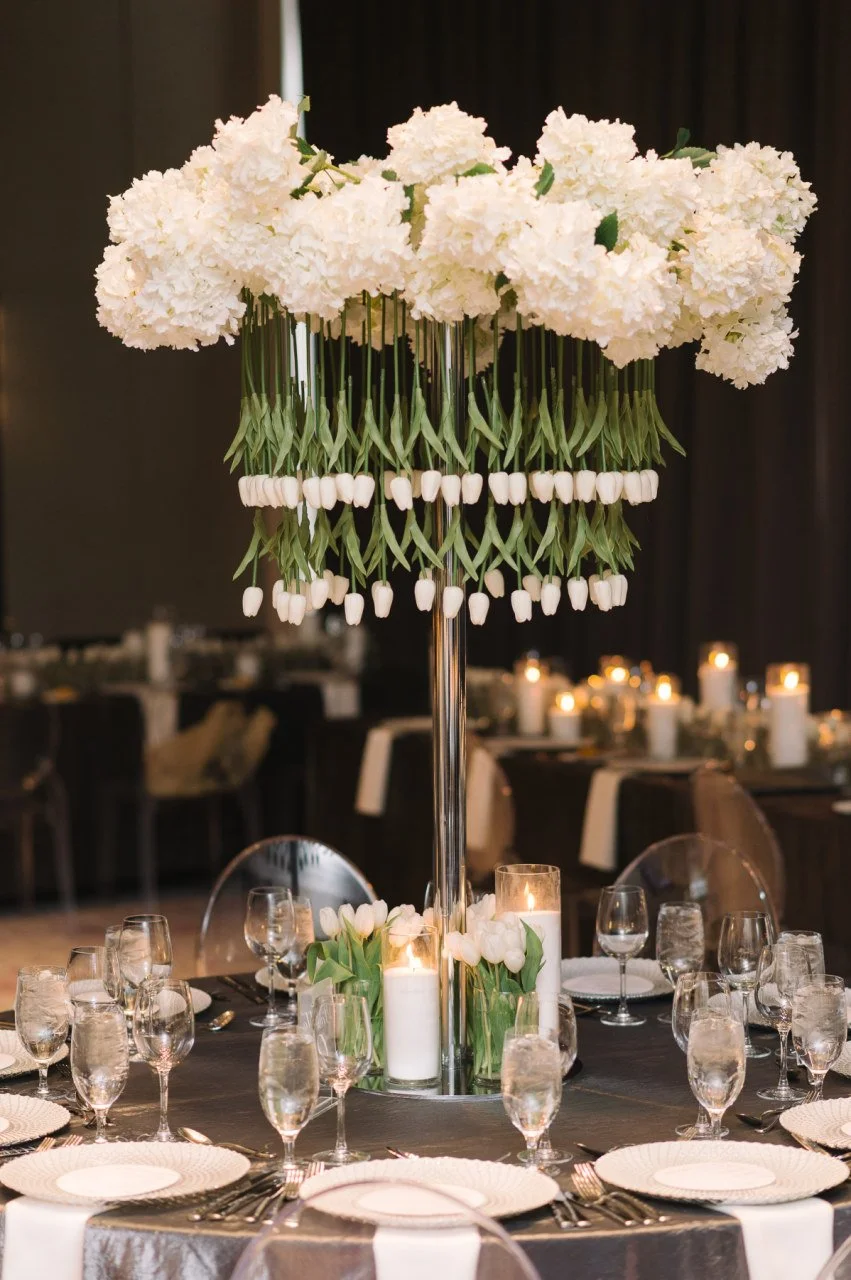 Elegant banquet table decoration with a tall floral centerpiece of white hydrangeas and tulips, surrounded by candles, set for a formal event.