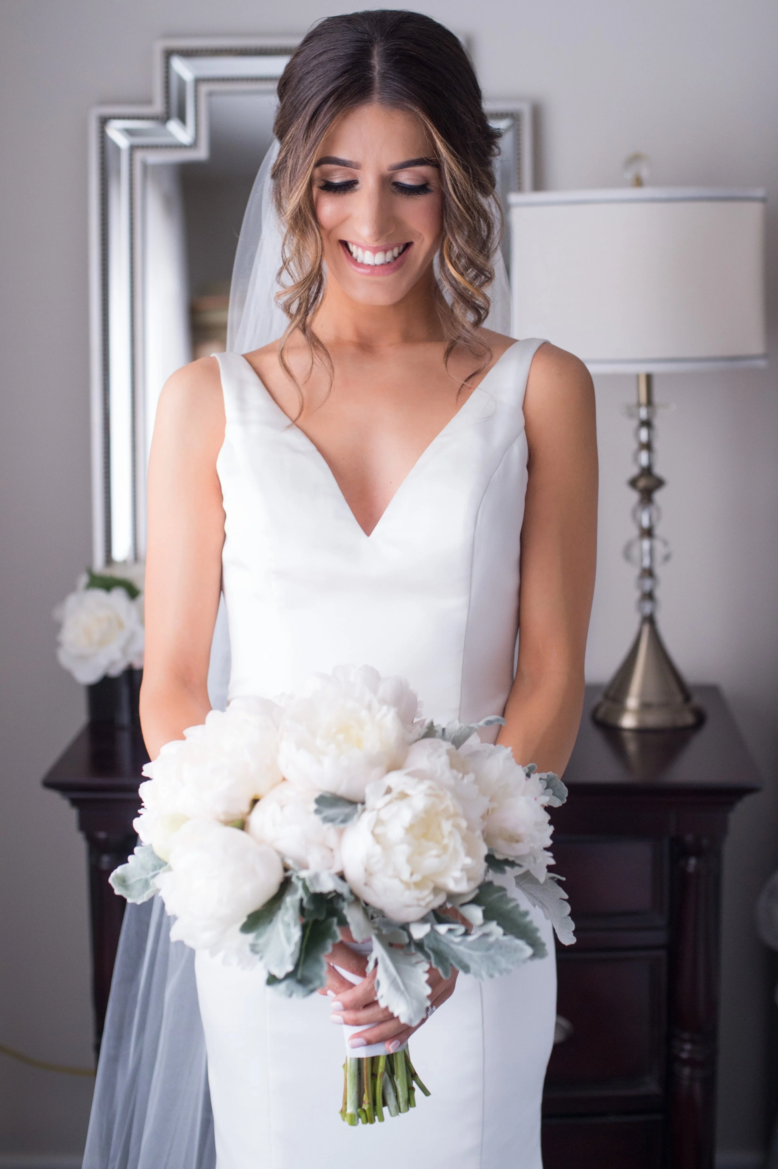 A smiling bride with wavy brown hair, wearing a white wedding dress, holding a bouquet of white flowers, in a room with a mirror and a dark wooden table with a white lamp in the background.