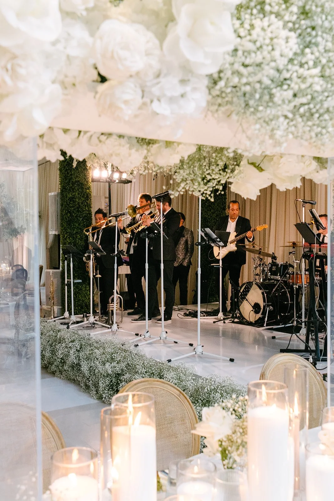 A band playing music on a stage at a wedding reception, with floral decorations overhead and table settings with candles in the foreground.