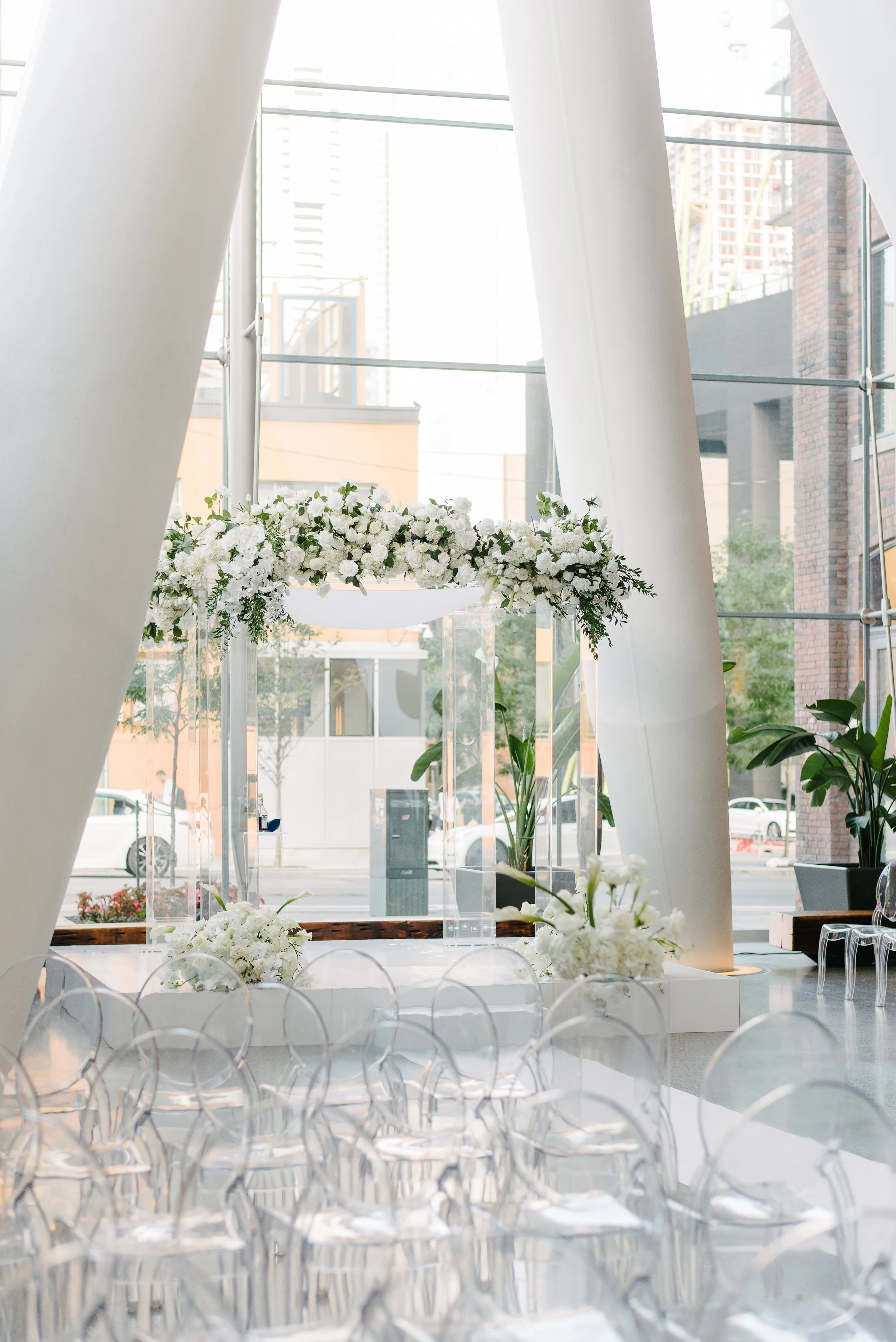 Interior of a modern venue with white drapery, floral arrangements, and clear chairs, set for an event like a wedding or reception, with large windows and city buildings visible outside.