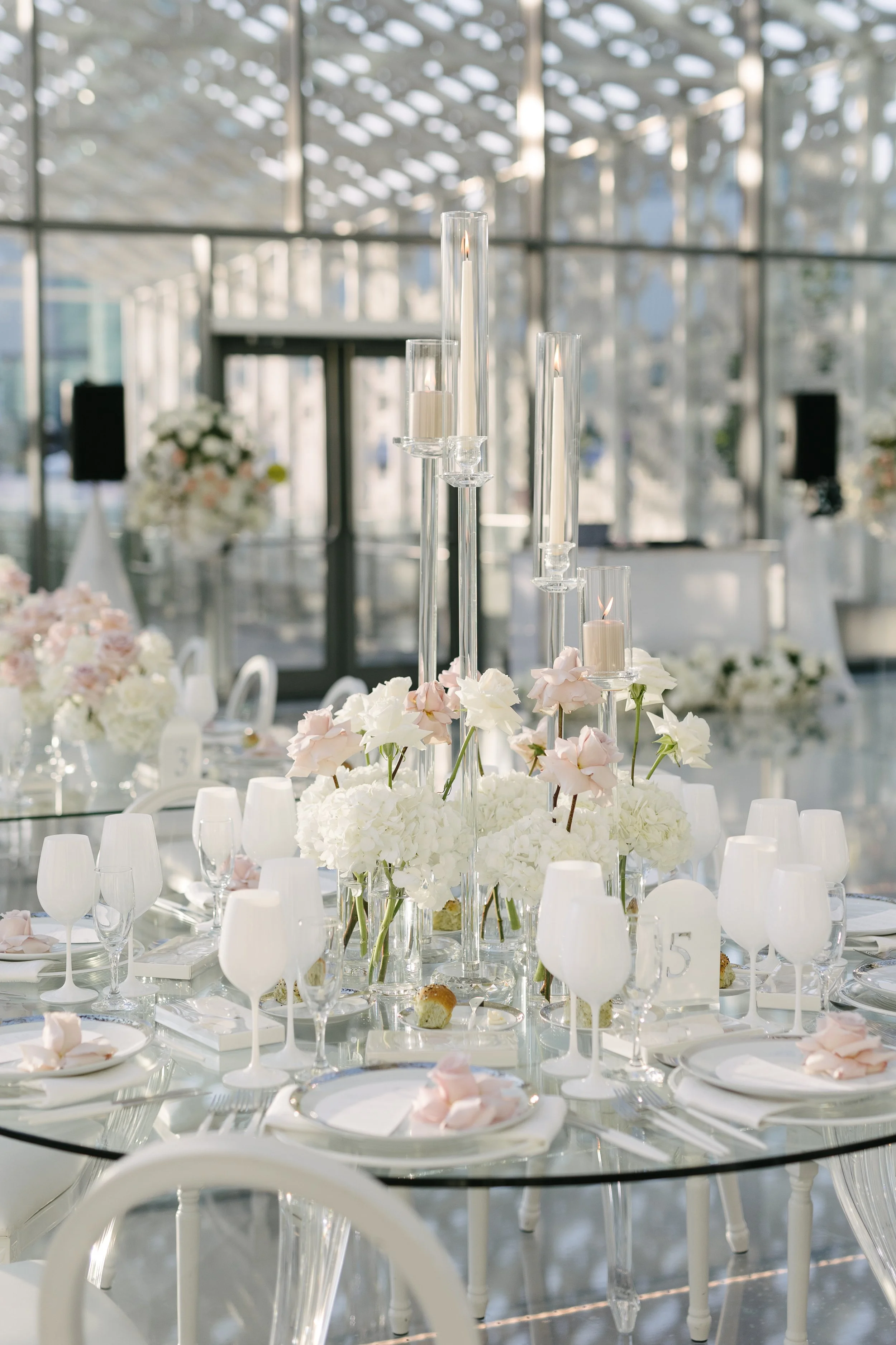 Elegant wedding reception table with white and blush pink floral arrangements, tall glass candle holders with lit white candles, white plates, glassware, and white chairs in a bright venue with large windows.