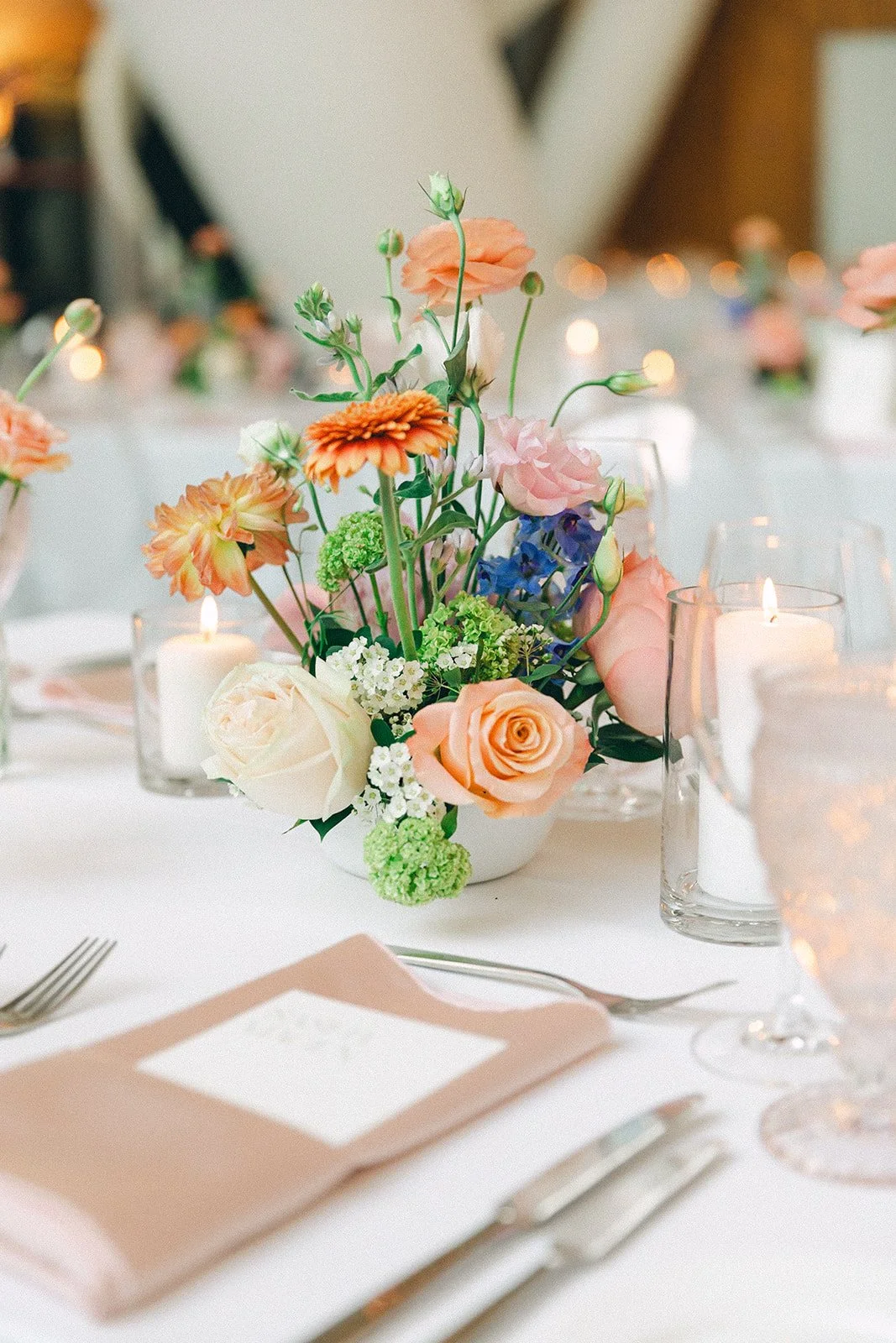 Flower arrangement with roses, ranunculus, and other flowers on a decorated table with candles and place settings at a formal event.