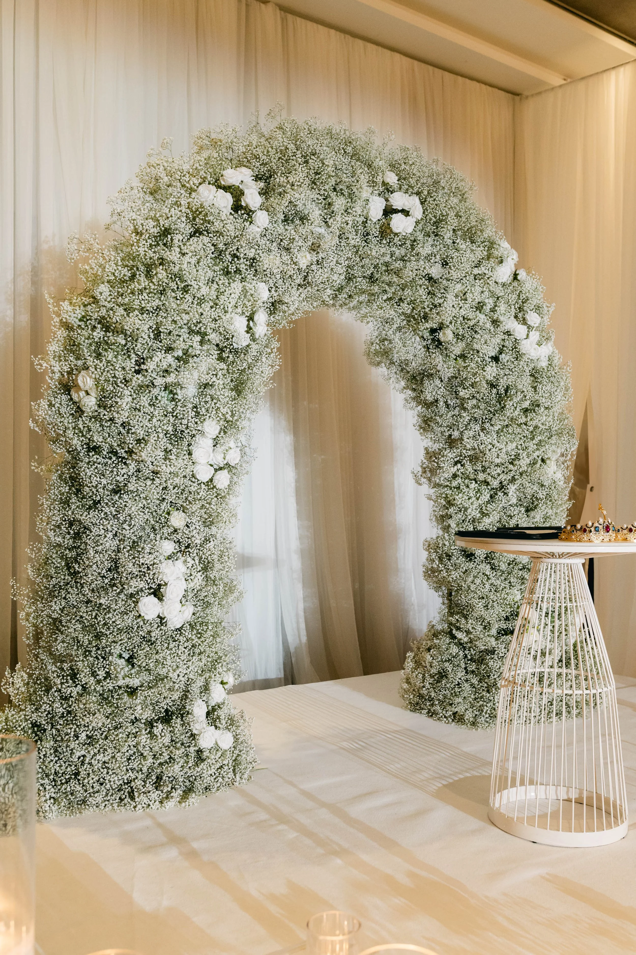 White floral arch made of baby’s breath and roses, with a small table and decorative cage at a wedding venue.