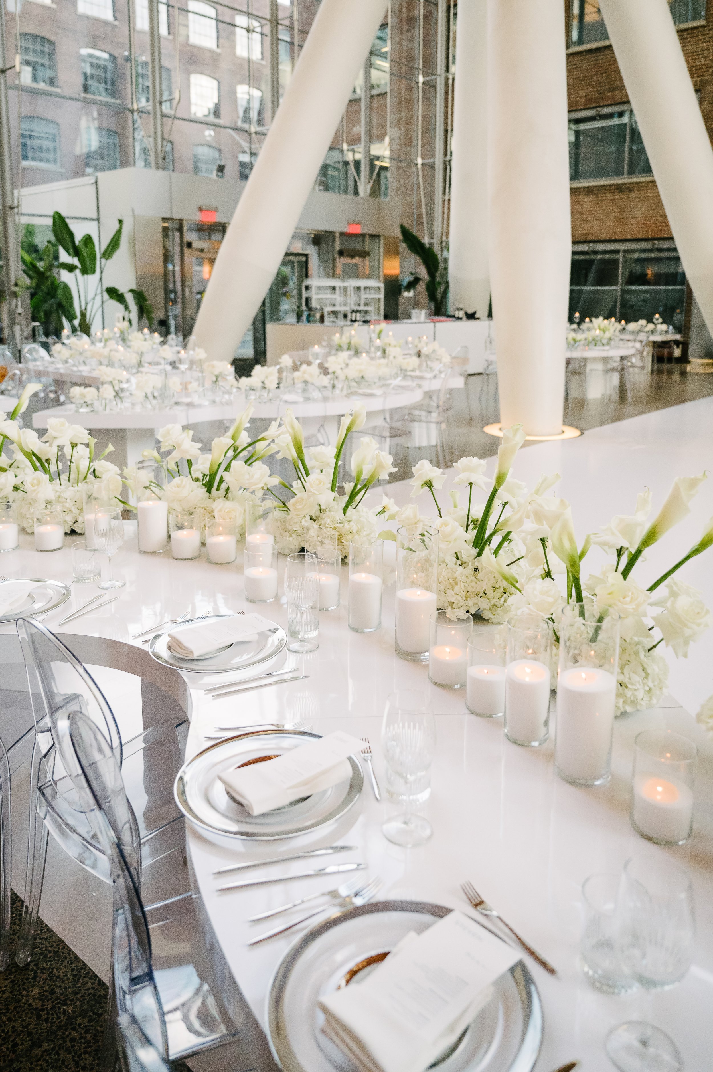 Elegant banquet table set with white floral arrangements, candles, glassware, and silverware in a modern enclosed space with large windows and exposed brick walls.
