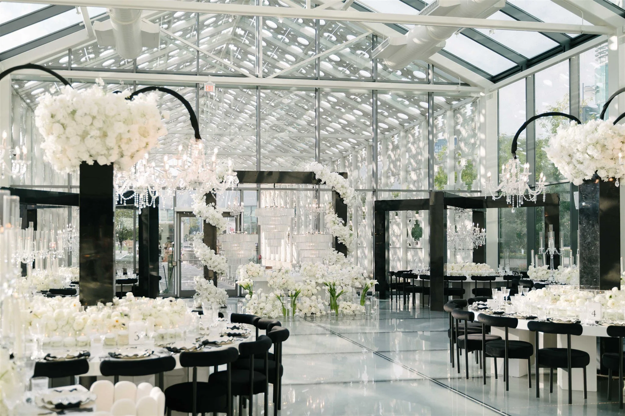 A bright, modern event space with glass walls and ceiling filled with white floral arrangements and chandeliers, set up for a wedding or celebration.