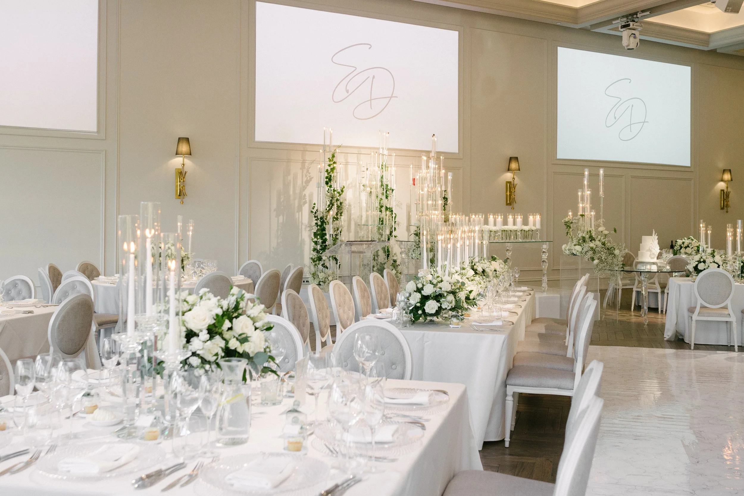 Elegant wedding reception hall with round tables covered in white tablecloths, decorated with white floral centerpieces and lit candles. The background features tall candle arrangements with greenery on a glass table, and large screens on the wall di