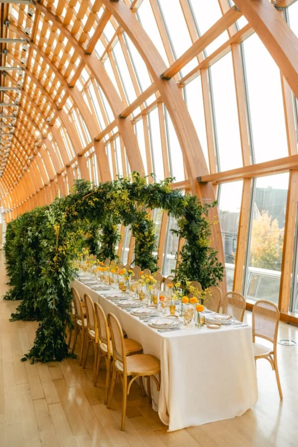 Long banquet table decorated with yellow flowers and glassware in a wooden, glass-ceilinged venue with an arch of greenery overhead.