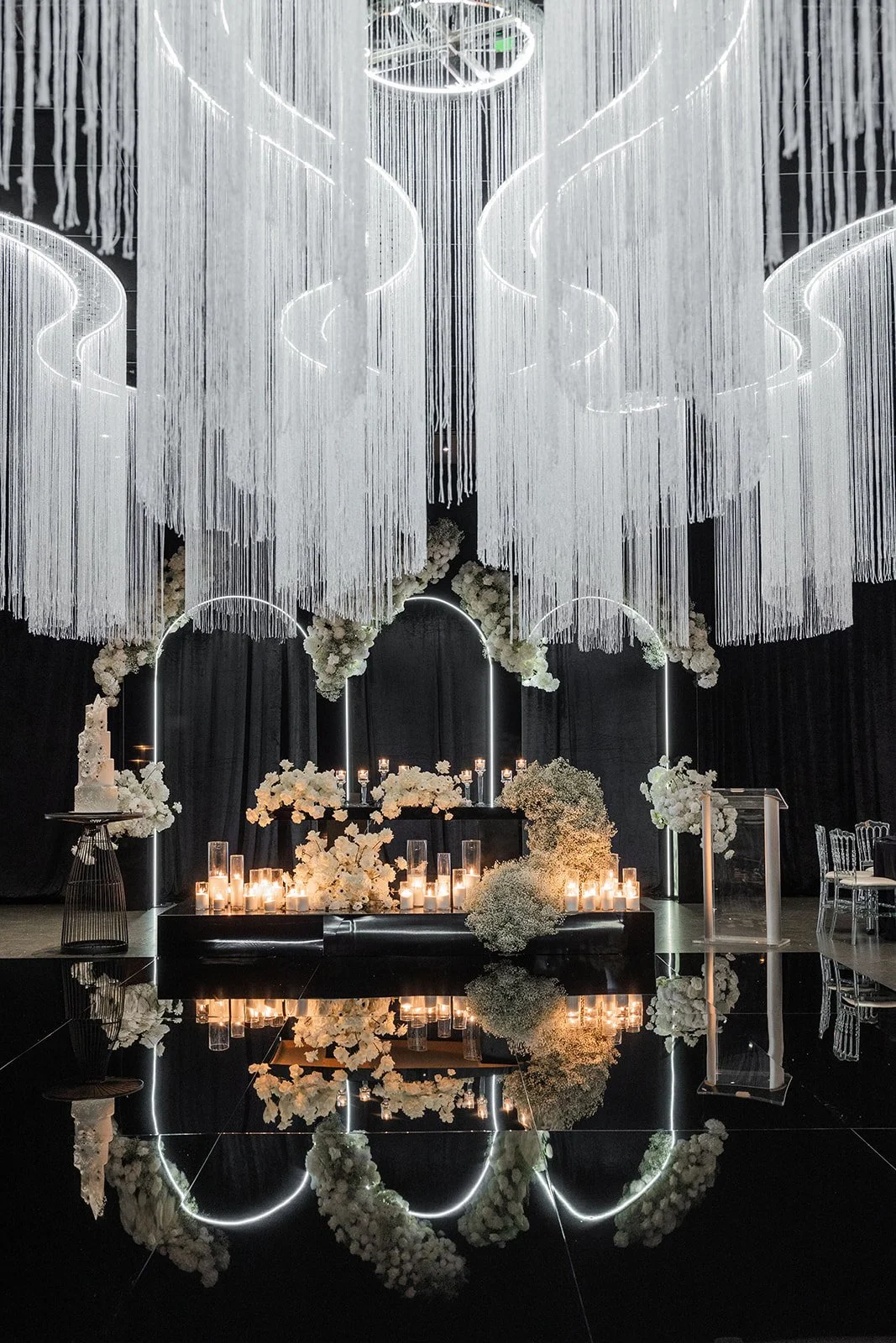 Elegant wedding or event altar with white flowers, candles, and modern light decorations on a black backdrop with a reflective black floor.