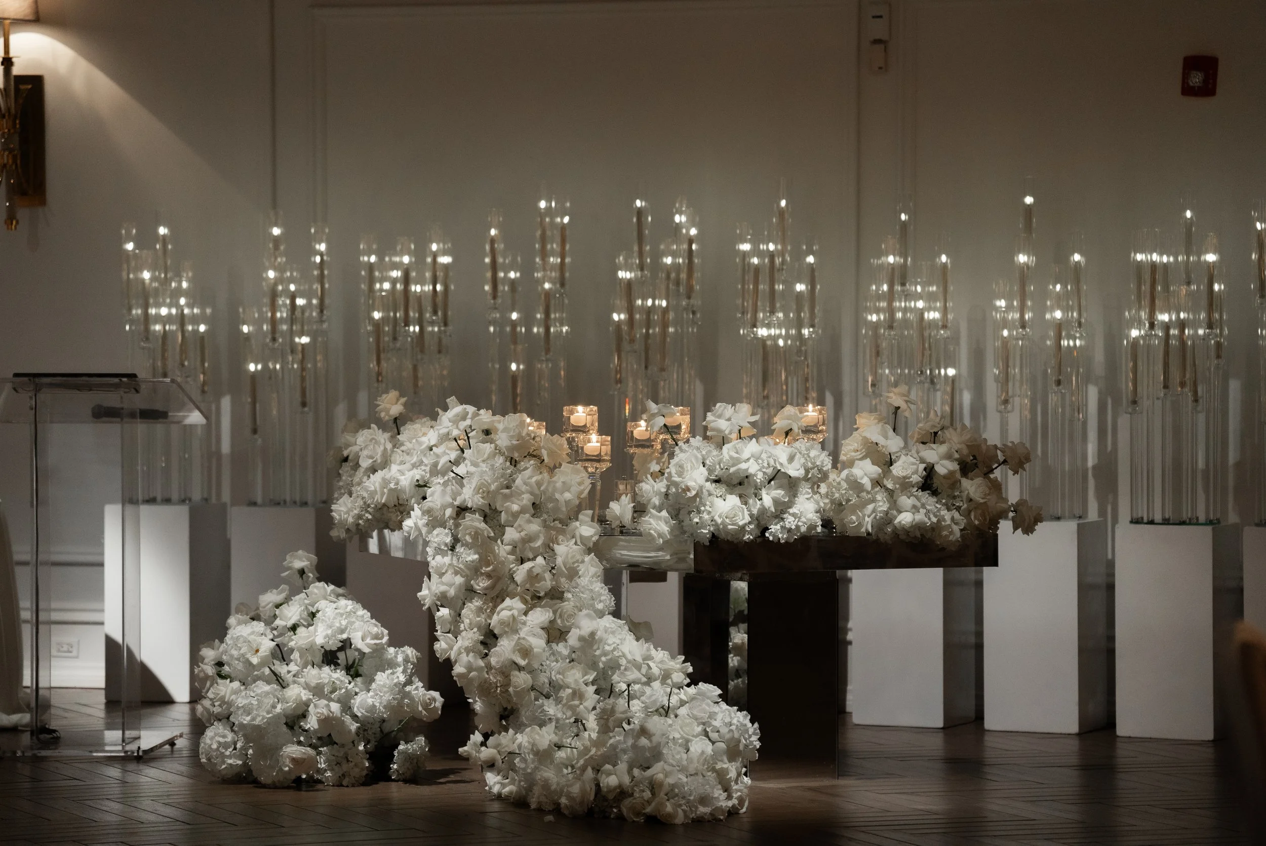 Indoor scene with a table or platform covered in white floral arrangements and candles, with hanging glass candle holders in the background, illuminated softly.