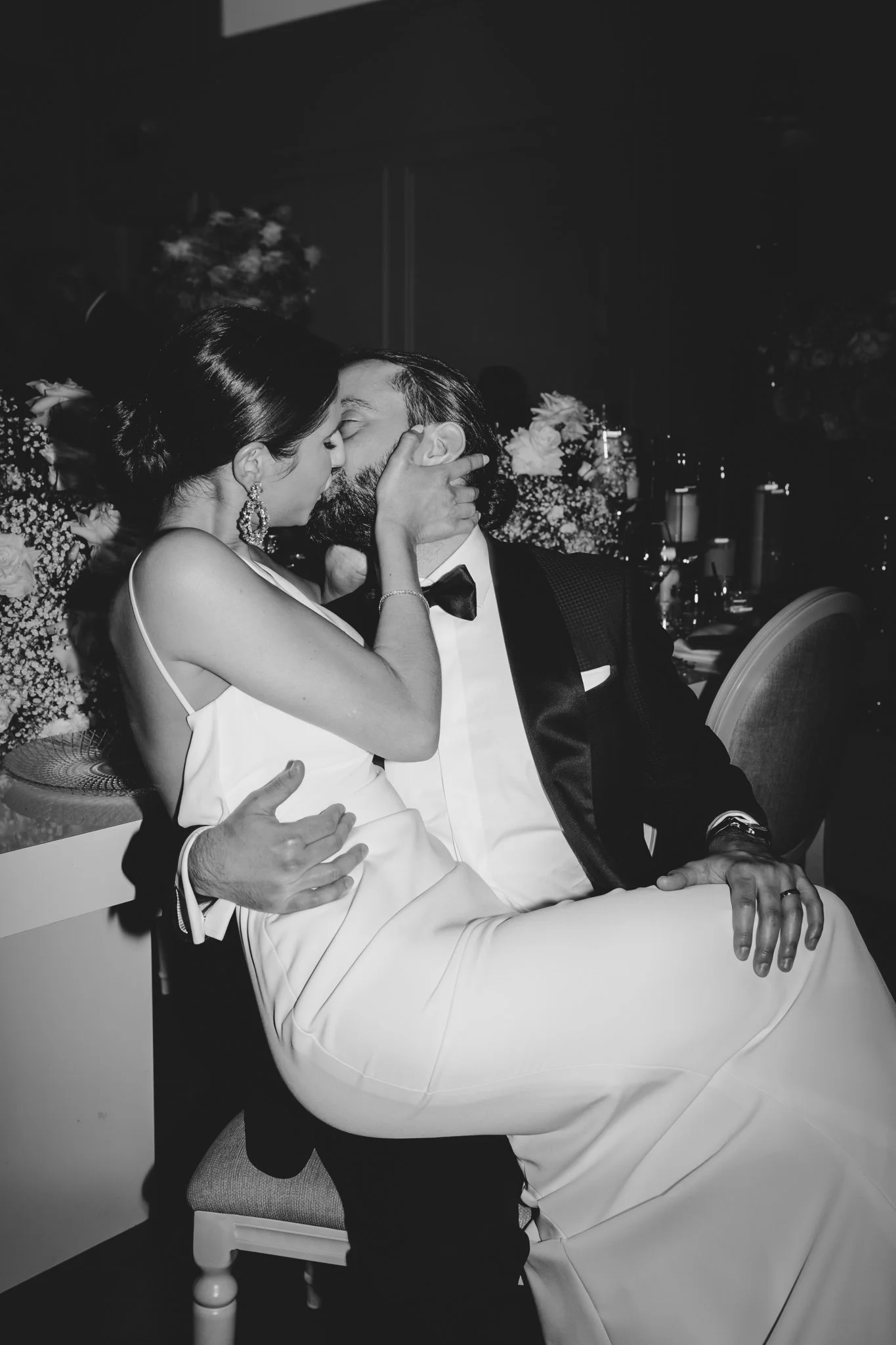 A black and white photo of a couple sharing a kiss at a formal event, dressed elegantly with flowers in the background.