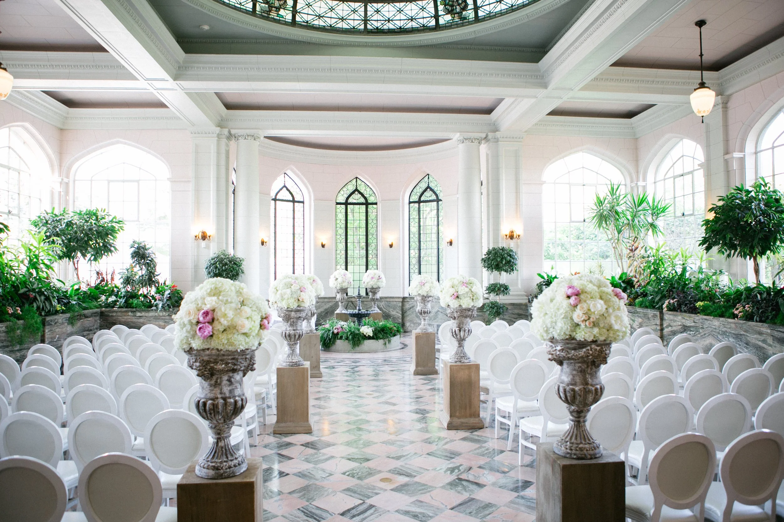Elegant indoor space with large arched windows, white walls, and floral arrangements on fountains and tables, set up for a wedding ceremony or event.