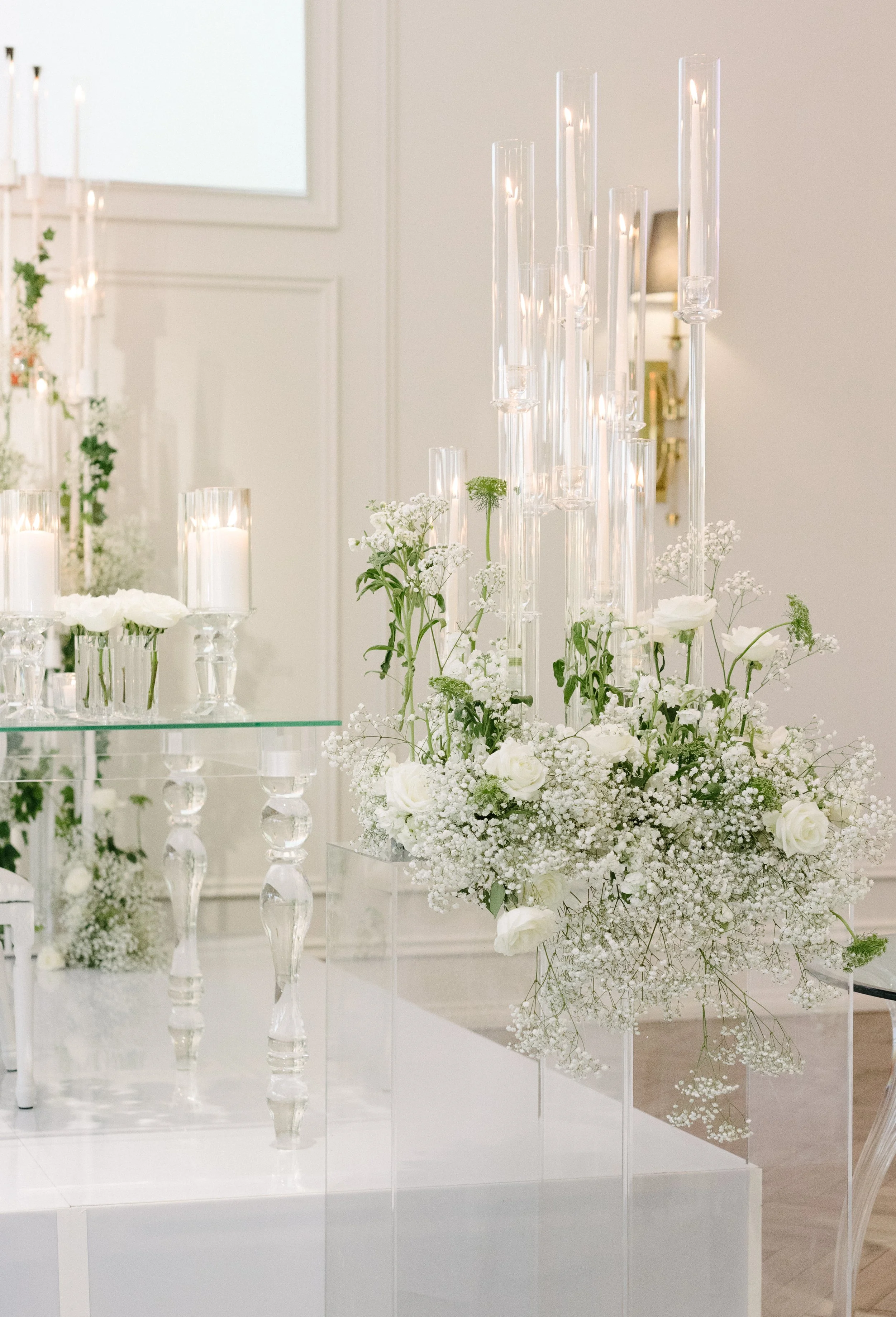 Elegant floral centerpiece featuring white roses, baby's breath, and greenery with tall glass candle holders and lit candles at a white event.