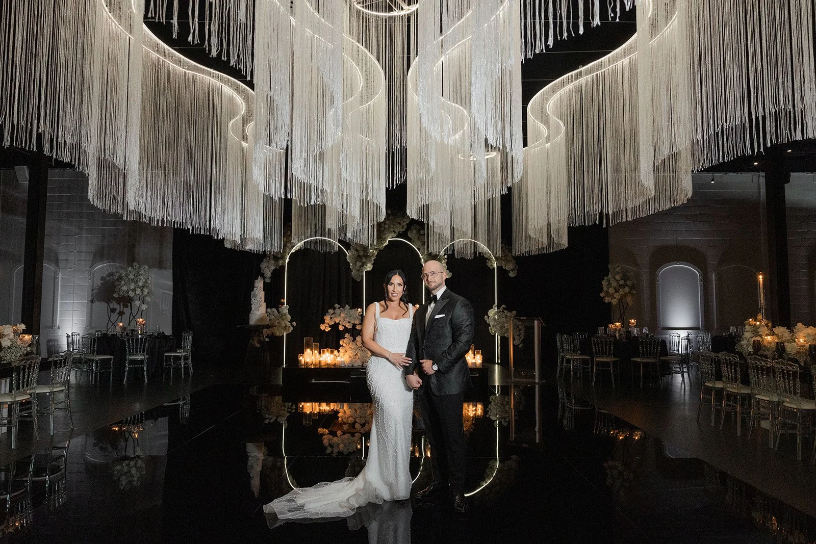 A bride and groom standing together at their wedding reception, decorated with elegant white floral arrangements, candles, and modern lighting fixtures in a dimly lit venue.