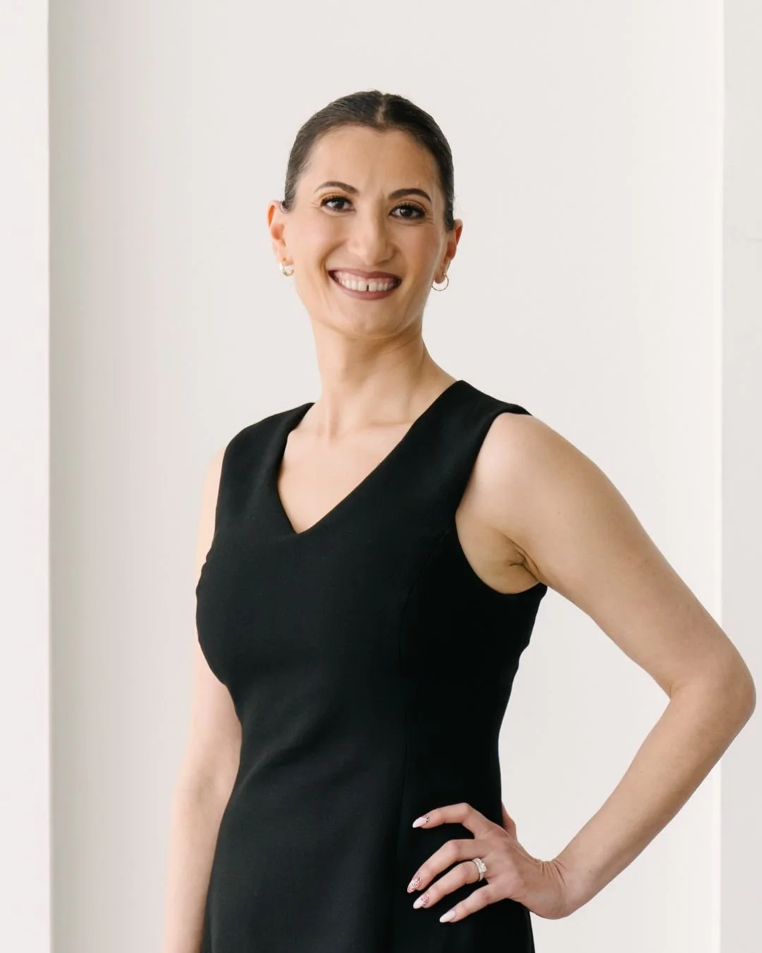A smiling woman with dark hair in a bun, wearing a sleeveless black dress, standing with one hand on her hip, in front of a white background.