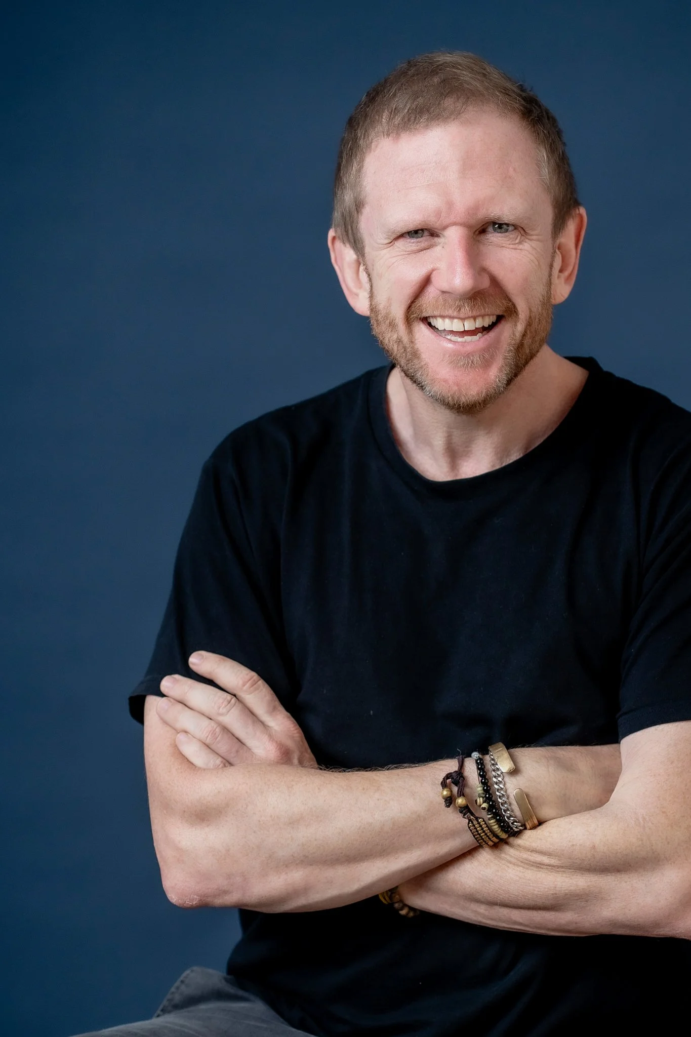 A smiling man with short, light brown hair and a beard, wearing a black T-shirt, bracelets on his right wrist, standing against a solid dark blue background.