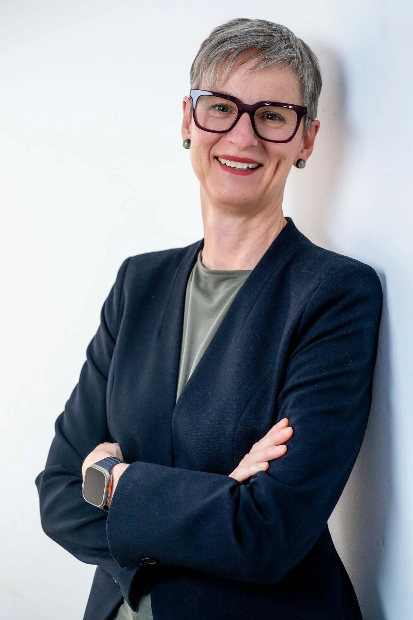 A smiling woman with short gray hair, wearing glasses, a black blazer, and a gray top. She has earrings, a smartwatch on her left wrist, and is standing with arms crossed against a plain white background.