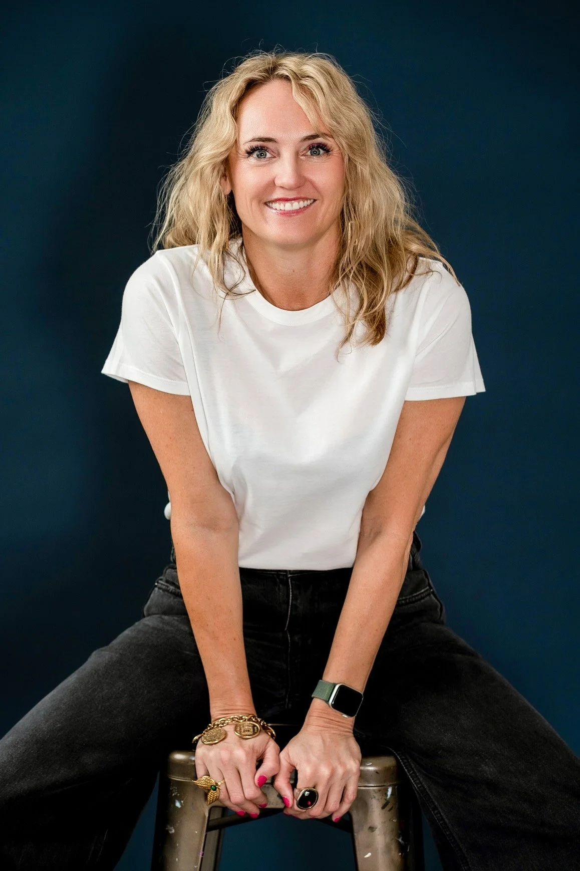 A woman with blonde wavy hair wearing a white t-shirt, black jeans, and jewelry, smiling while sitting on a stool against a dark blue background.