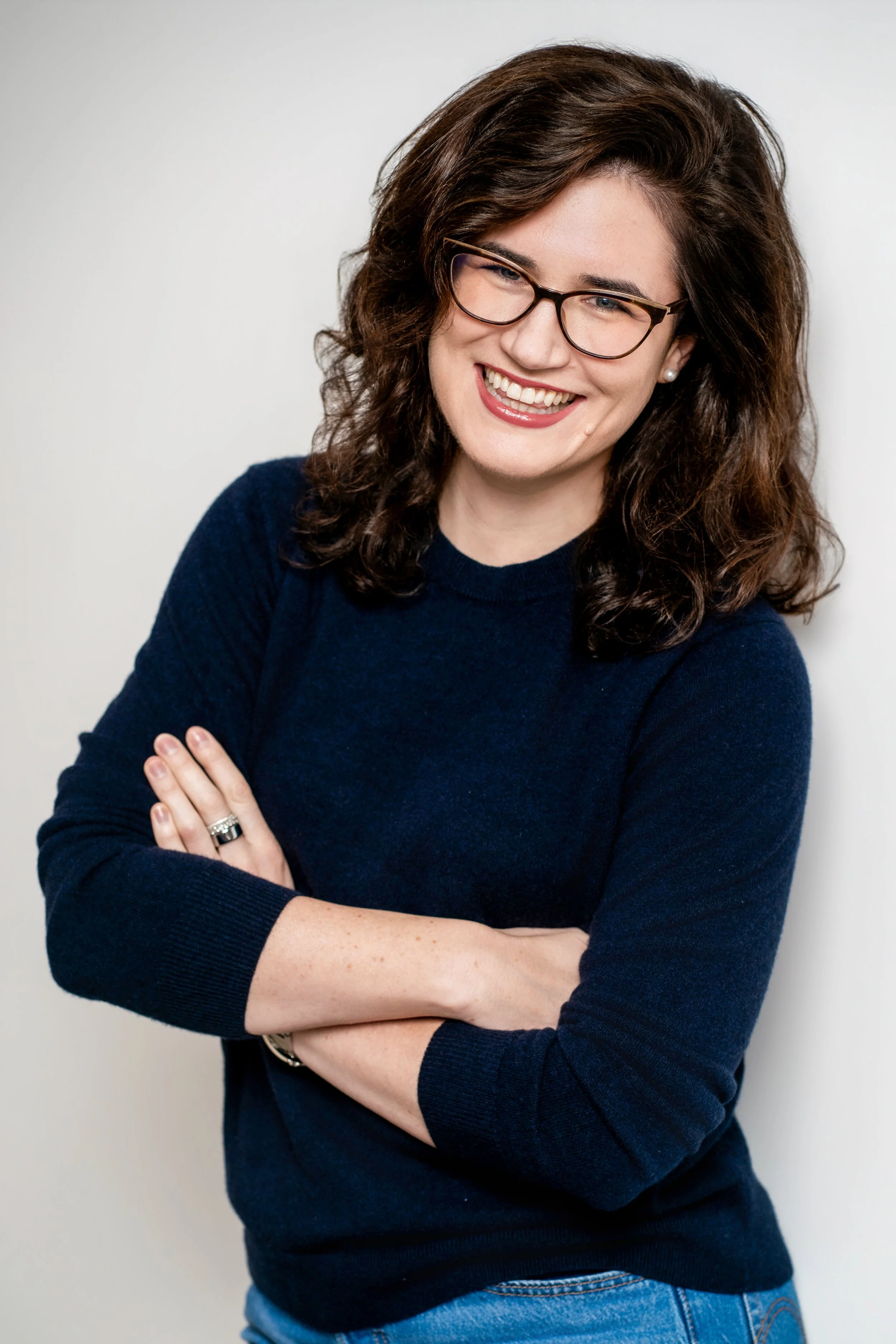 A woman with curly brown hair, wearing glasses, a navy blue sweater, and blue jeans, smiling with crossed arms against a plain white background.