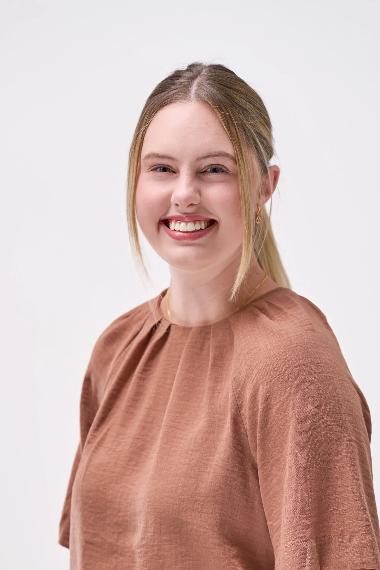 A young woman with blonde hair styled half-up, half-down, smiling and winking in front of a plain white background, wearing a brown textured top, small earrings, and a delicate necklace.