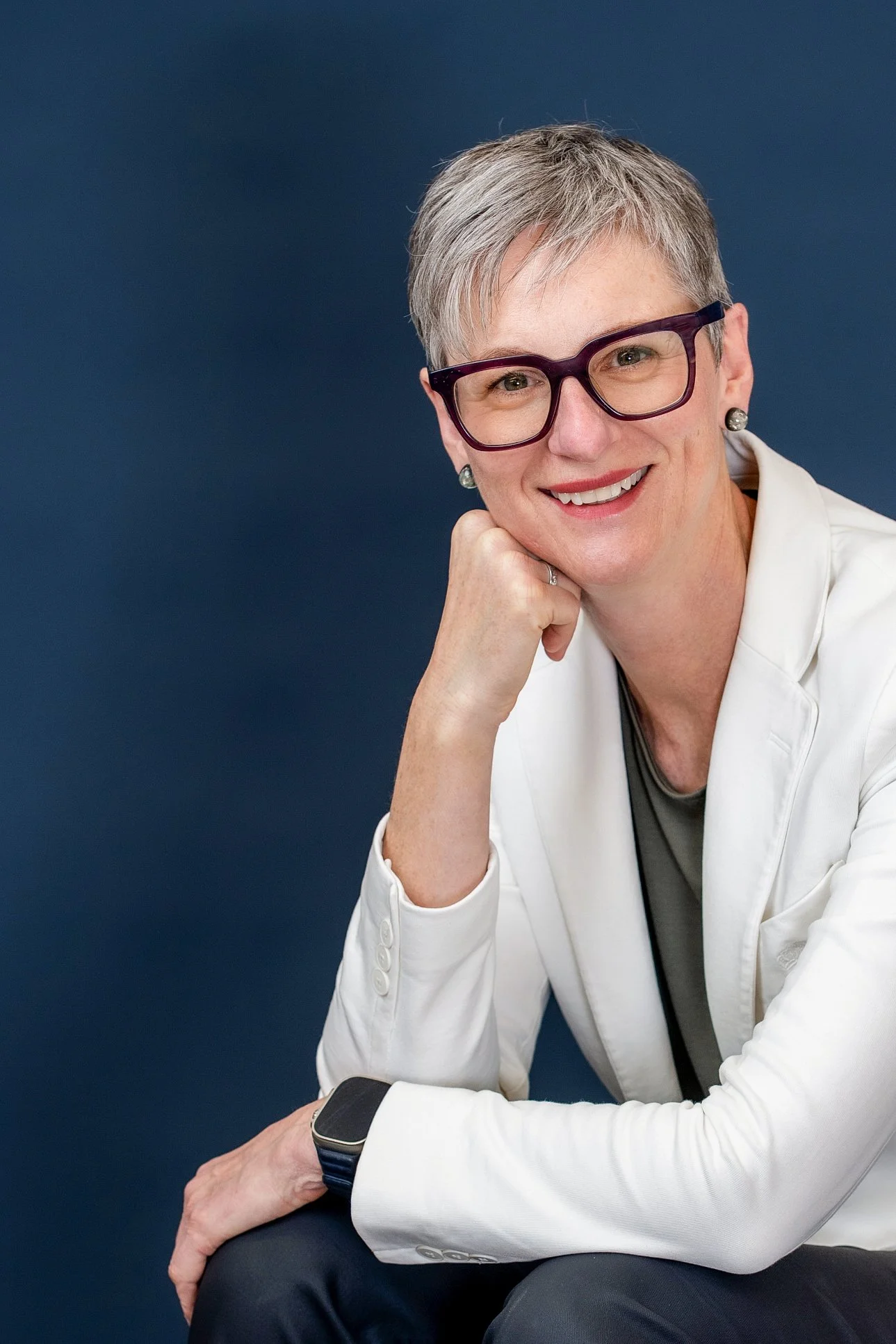 A middle-aged woman with short gray hair, glasses, and earrings, wearing a white blazer, smiling and sitting at a table with a dark background.