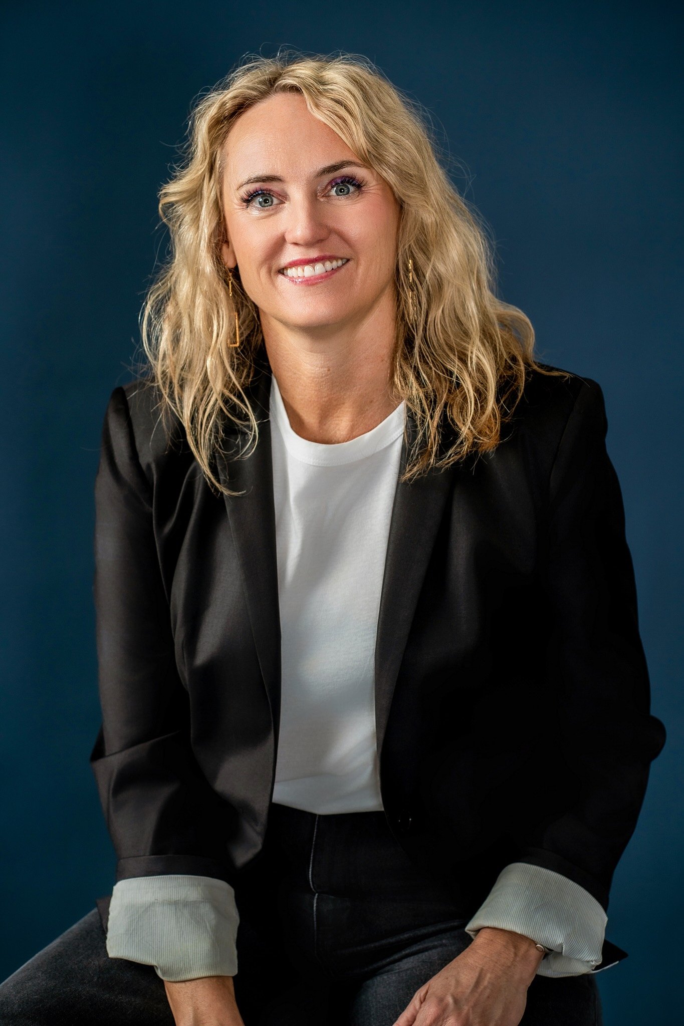 A woman with blonde, curly hair, wearing a black blazer over a white shirt, sitting against a blue background, smiling at the camera.