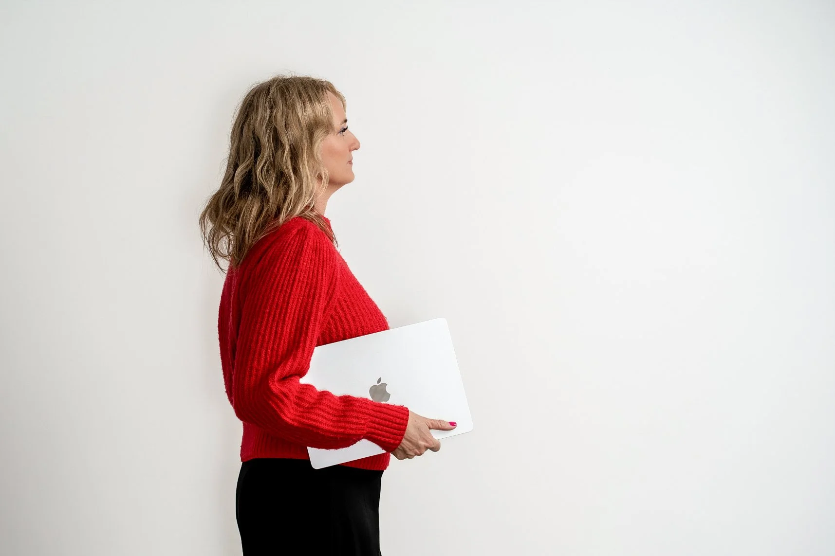 A woman with wavy blonde hair holding a closed silver MacBook laptop, standing in profile against a plain white wall, wearing a red sweater and black pants.