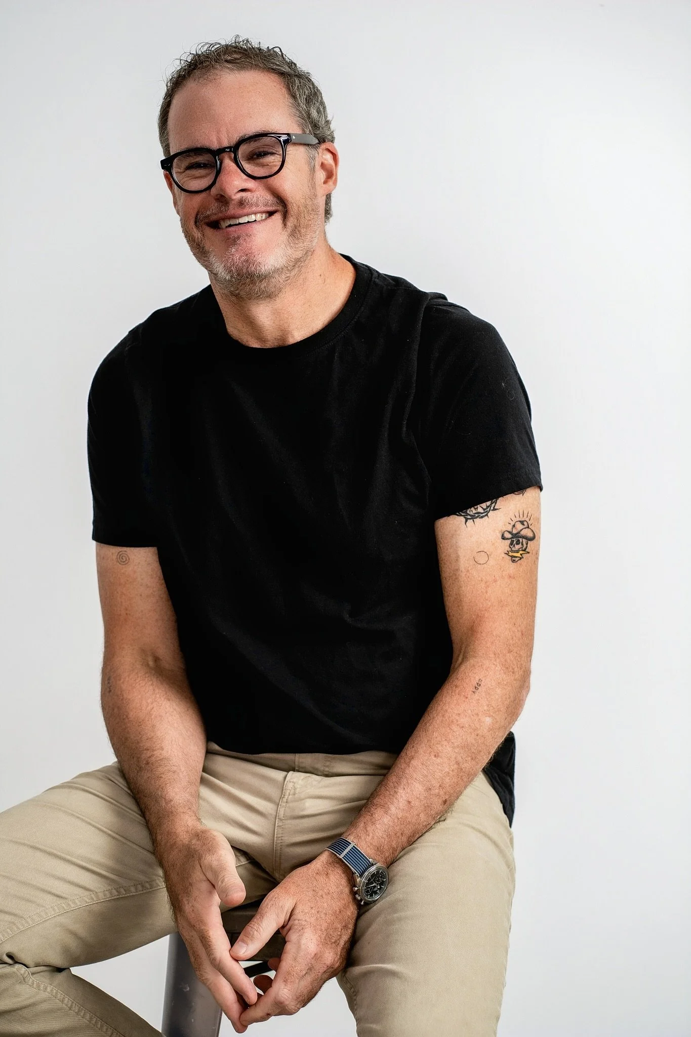 A smiling man with glasses wearing a black t-shirt and beige pants, sitting against a white background.