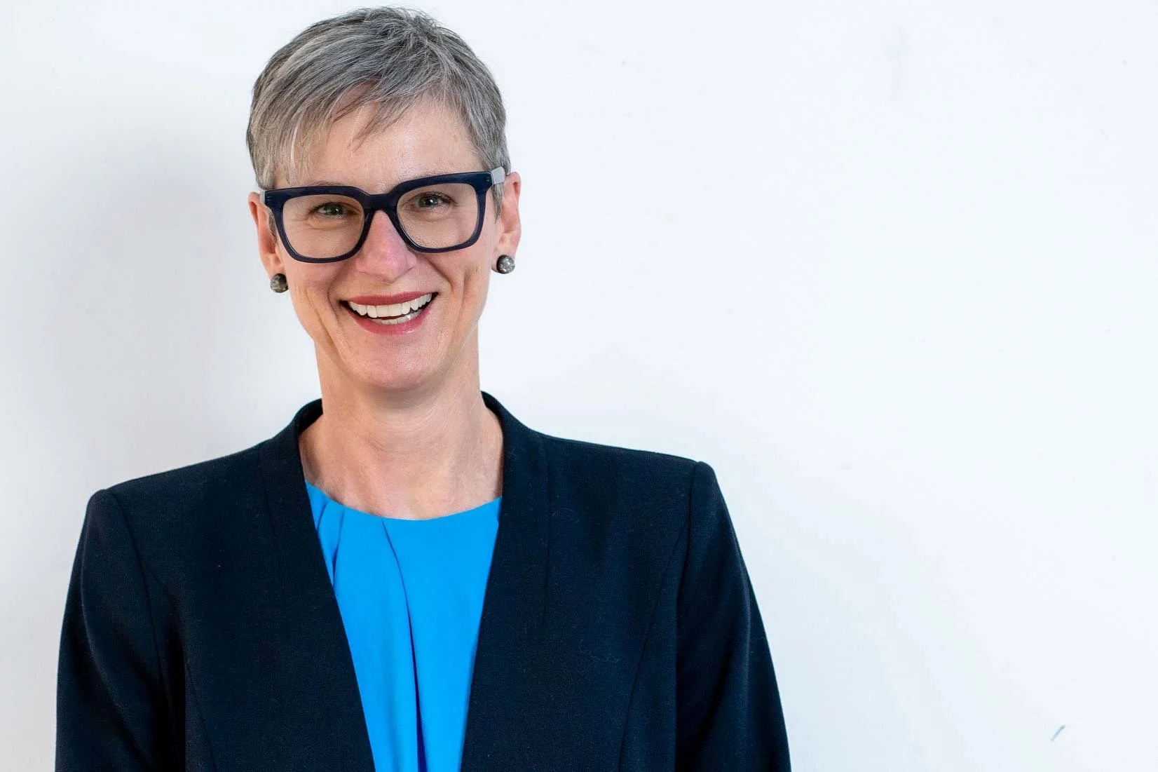 A smiling woman with short gray hair and black glasses, wearing a dark blazer and a bright blue top, standing against a plain white wall.