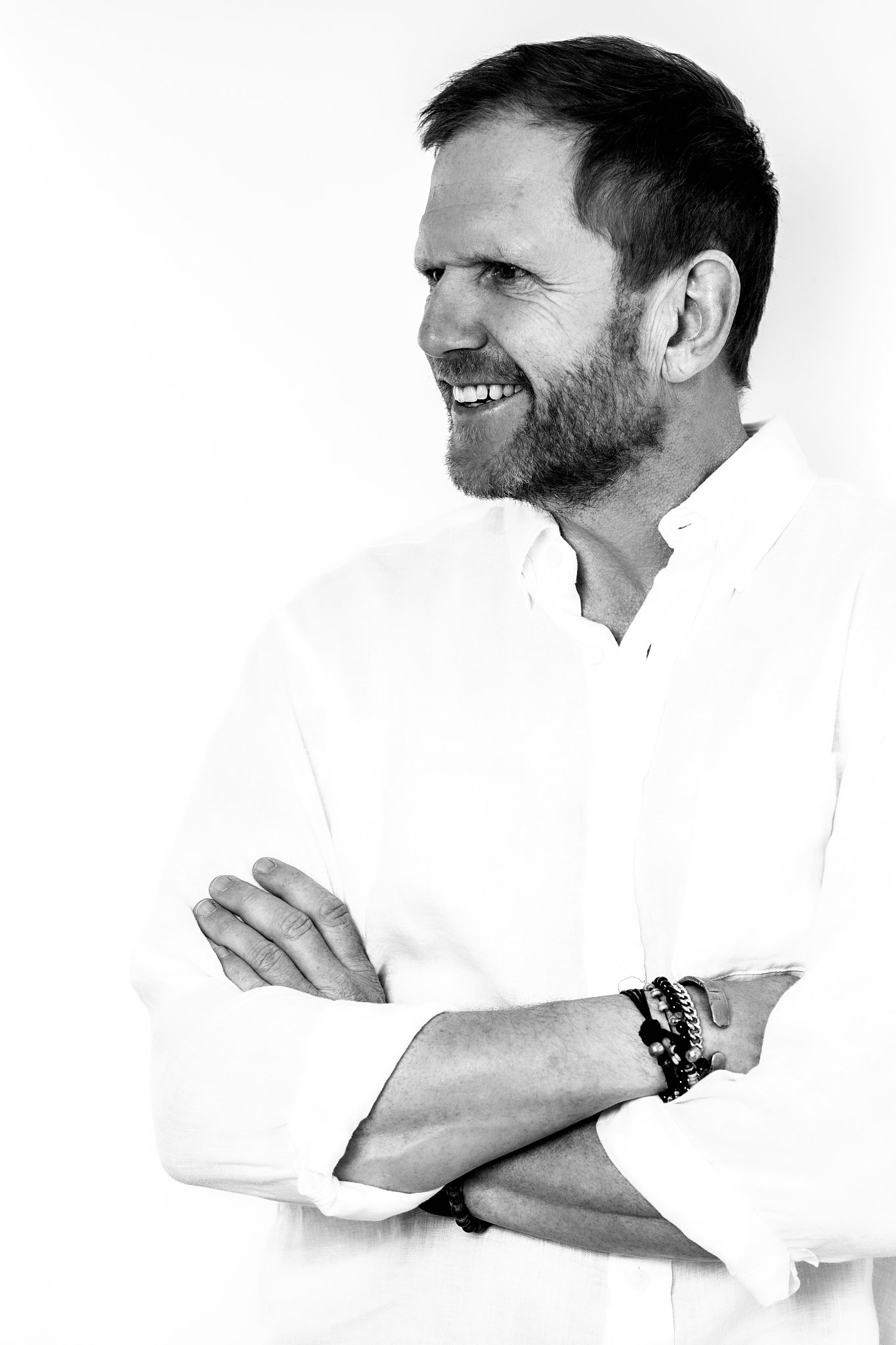 A black and white photo of a man smiling with his arms crossed, wearing a white shirt and bracelets on his wrist.