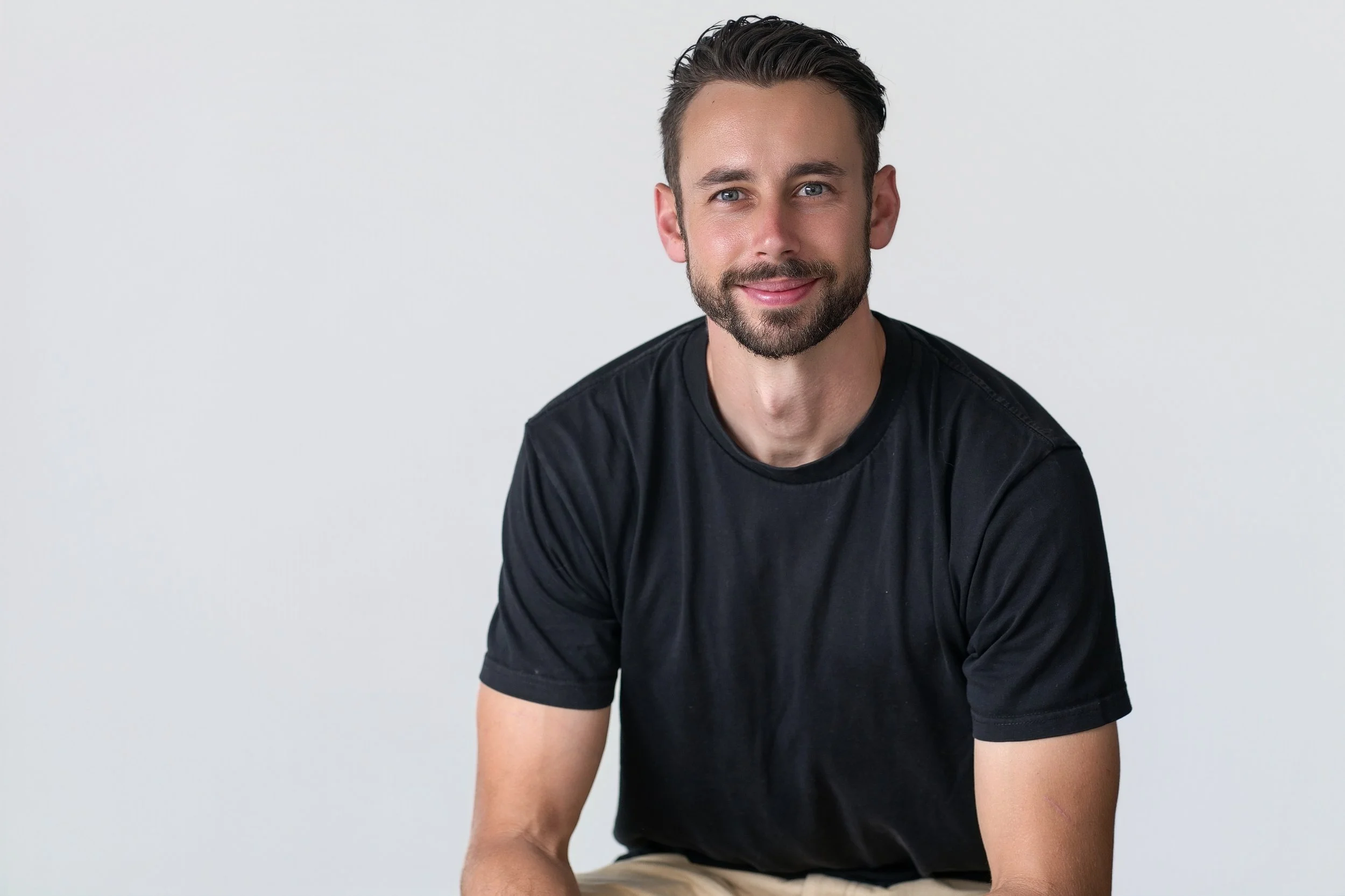 Portrait of a young man with dark hair and beard, wearing a black T-shirt, sitting against a plain light background.