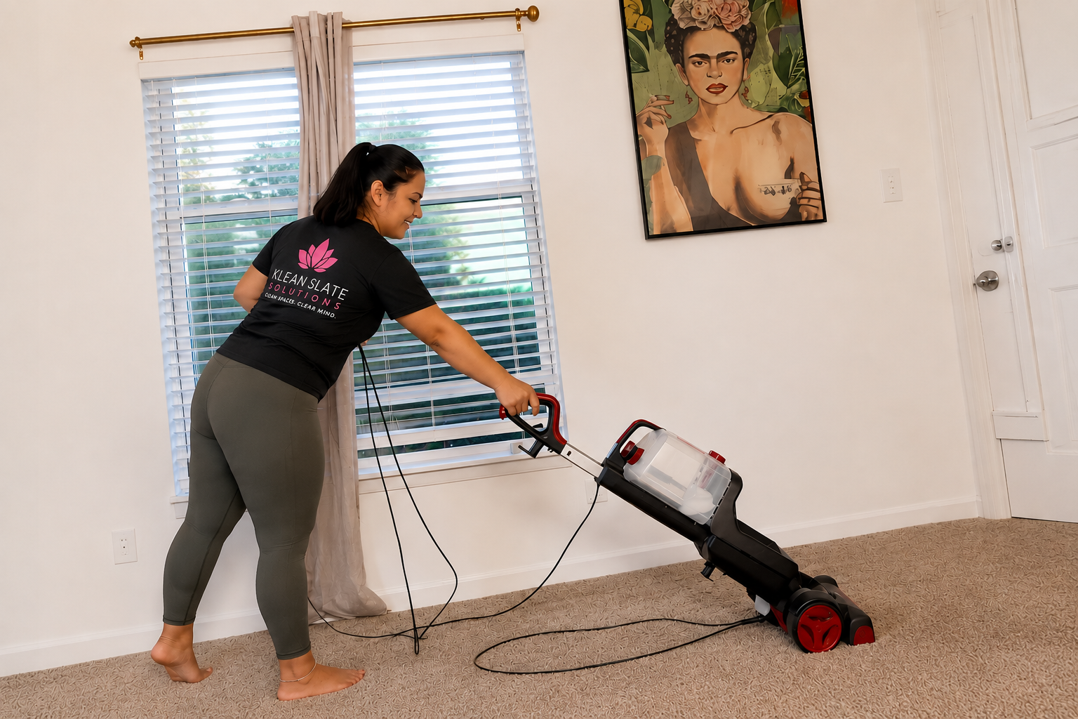 A woman vacuuming a beige carpet in a room with white walls and a large window has blinds and beige curtains. She is wearing a black t-shirt with pink and white text and gray leggings. Behind her, there is a colorful Frida Kahlo portrait hanging on the wall.