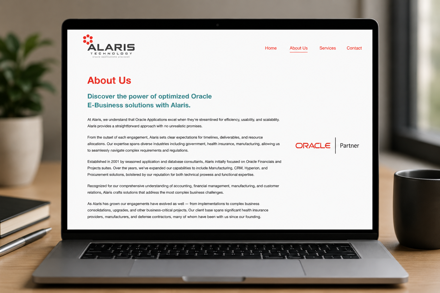 Laptop displaying the About Us page of Alaris Technology website with text about Oracle applications and company background, placed on a wooden desk with a black mug, pen, notebook, and potted plant nearby.