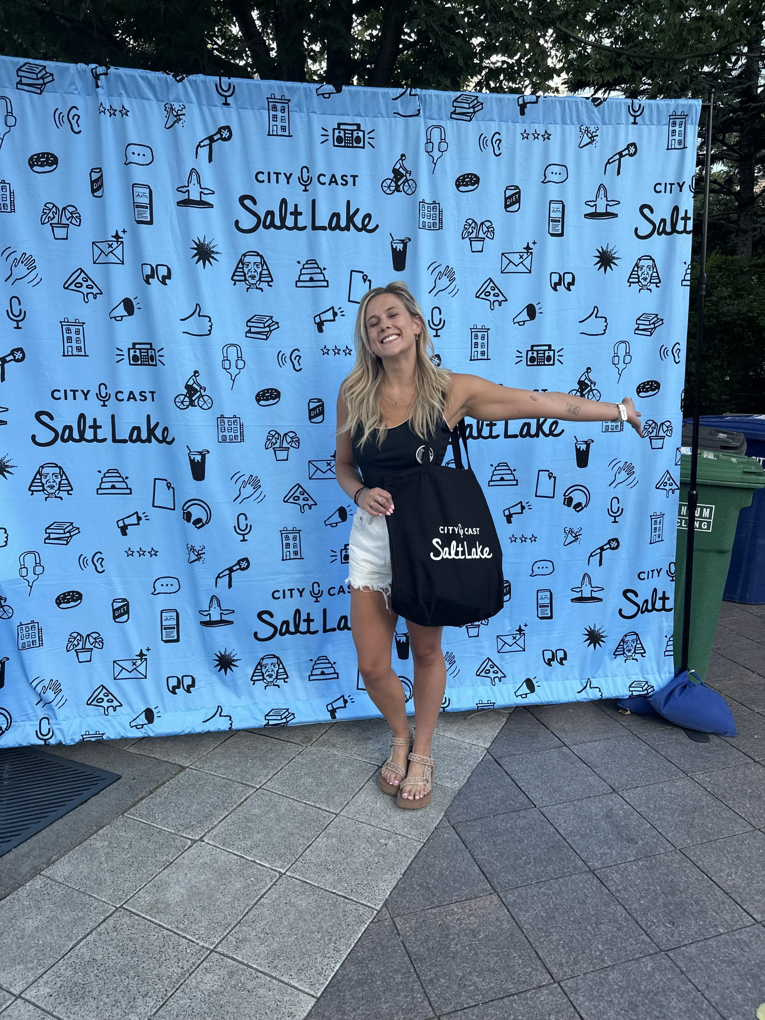 A woman standing outdoors in front of a blue backdrop with black line illustrations of various items such as books, a bicycle, a fountain, and speech bubbles. The backdrop has text that reads 'City Cast Salt Lake.' She is smiling with eyes closed, wearing a black sleeveless top, white shorts, and beige sandals, and is holding a black tote bag with the same logo.