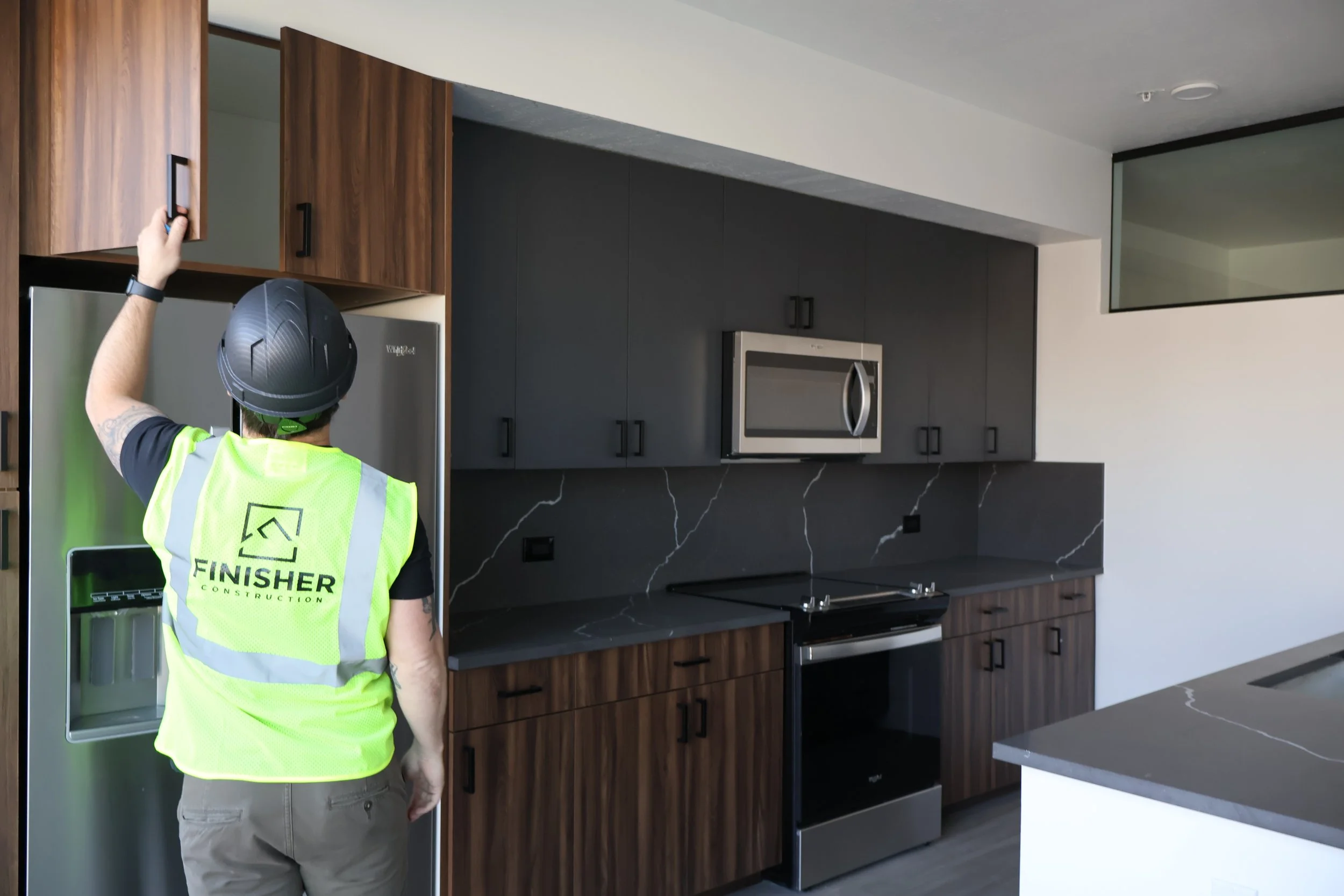 A construction worker in a neon yellow safety vest with 'FINISHER Construction' written on the back and a black helmet, facing away and reaching out to open a kitchen cabinet in a modern, dark grey and wood kitchen.
