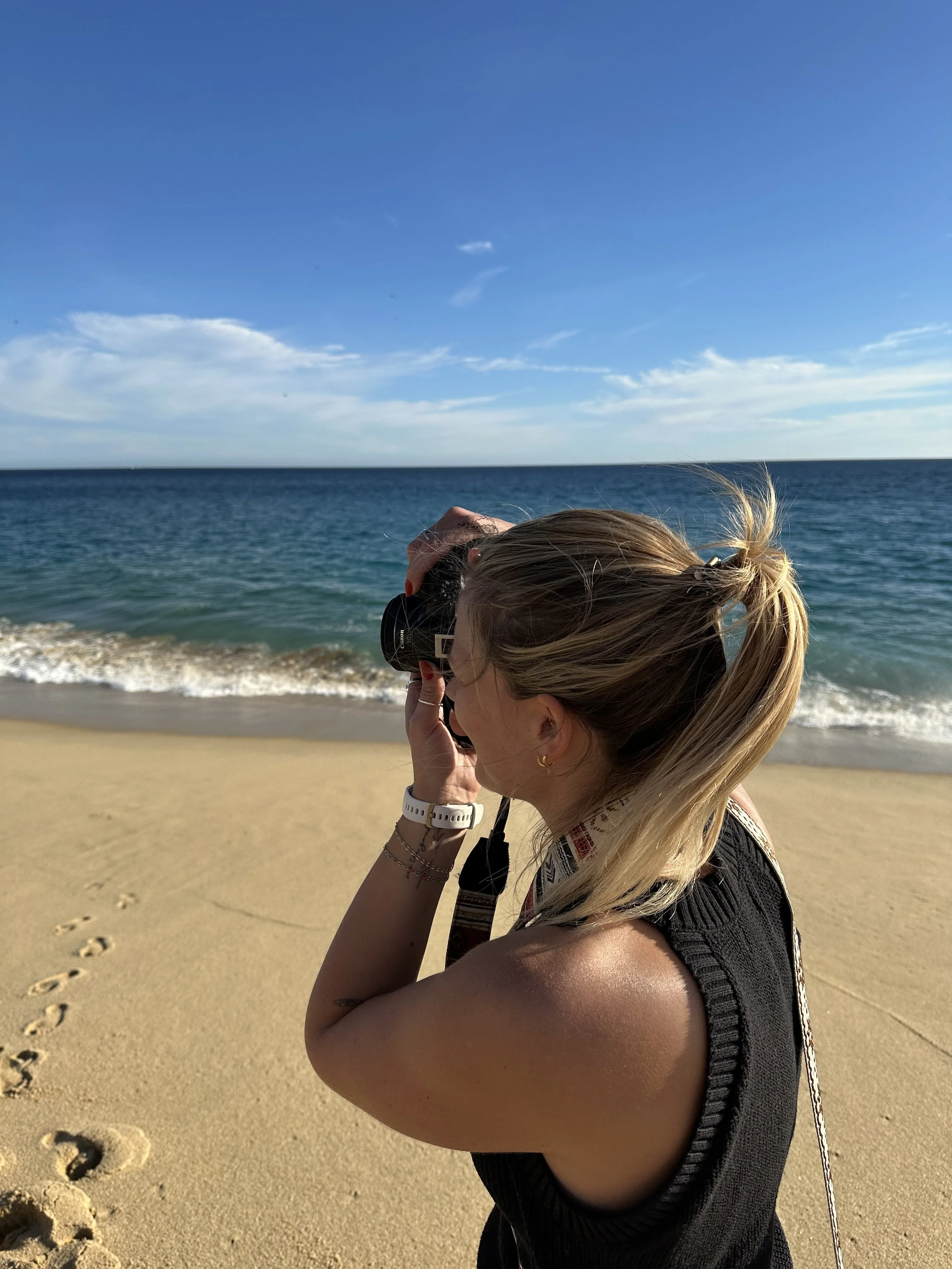 A woman with blonde hair tied in a messy bun, wearing a sleeveless black top, taking a photograph with a camera on a sandy beach with footprints, rolling waves, and a partly cloudy sky in the background.